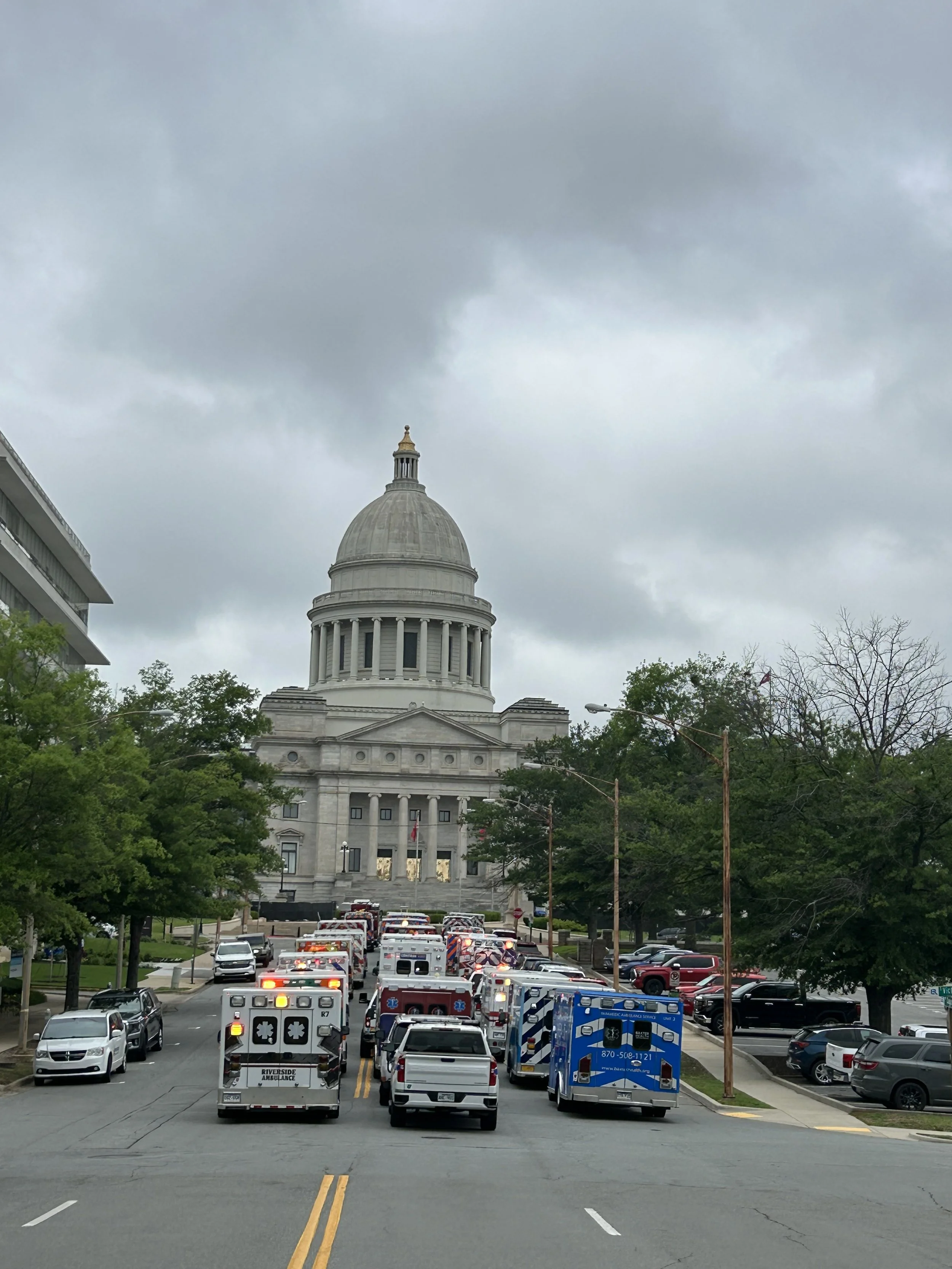 Street view with multiple emergency response vehicles, including ambulances, heading towards the state capitol building with a dome, under a cloudy sky.