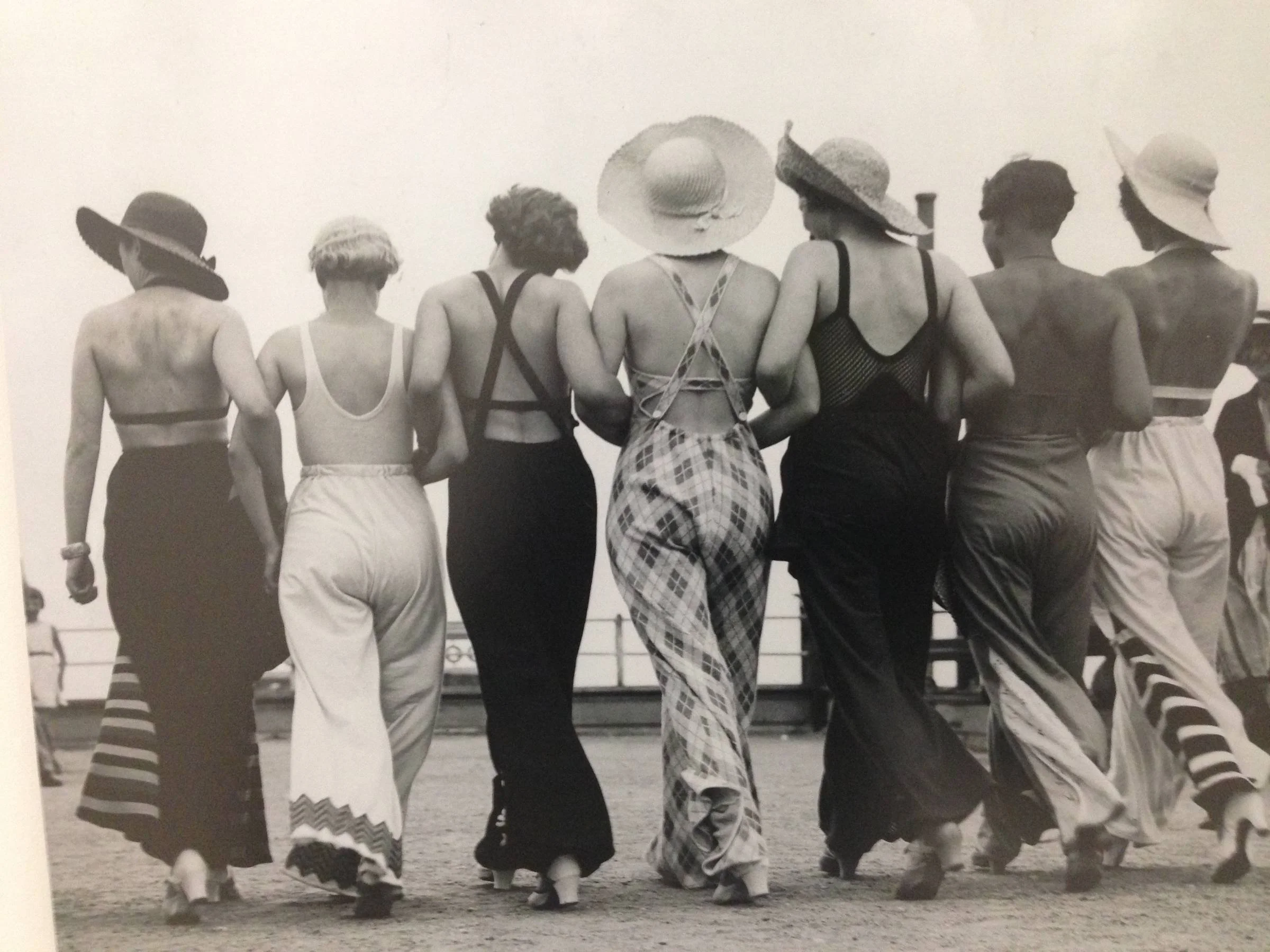 Black and white photo of seven women dressed in vintage bathing suits and wide-brimmed hats, walking on a beach or boardwalk.