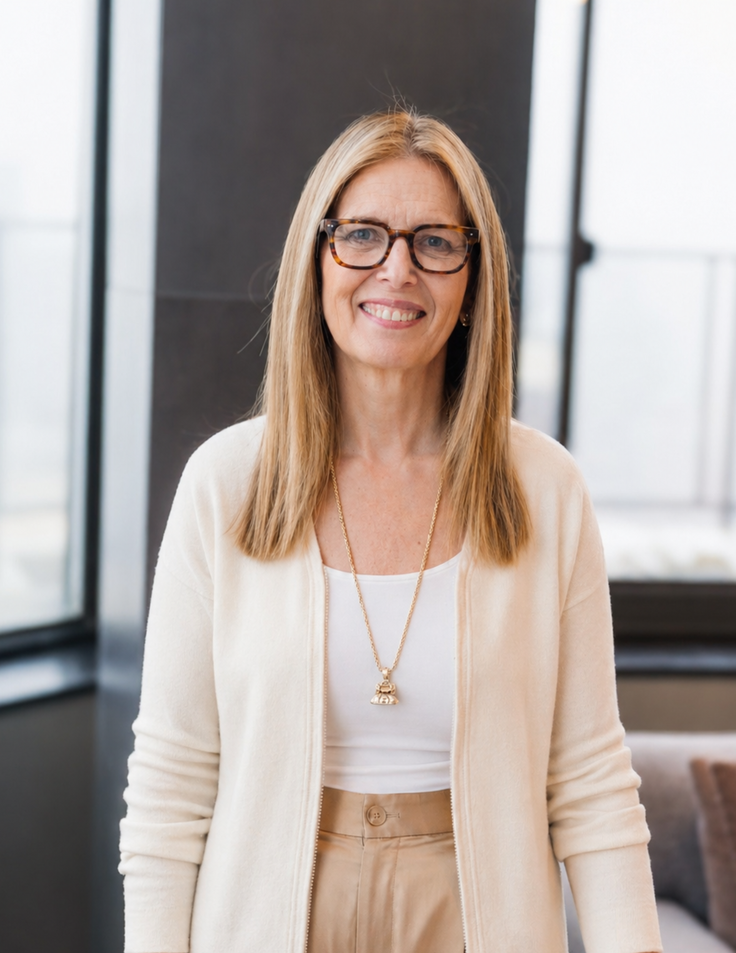 A middle-aged woman with blonde hair, wearing tortoiseshell glasses, a white top, a cream cardigan, and beige pants, smiling in an indoor setting with large windows in the background.