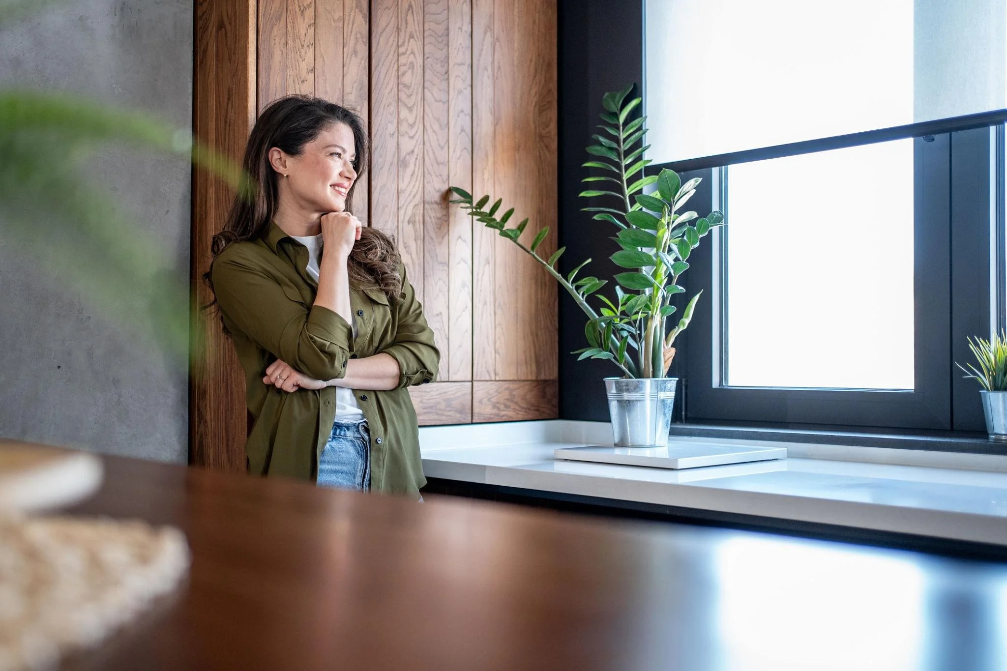 A woman with long dark hair smiling and leaning against a wooden wall in a modern kitchen, with plants on the counter and a window allowing natural light to enter.