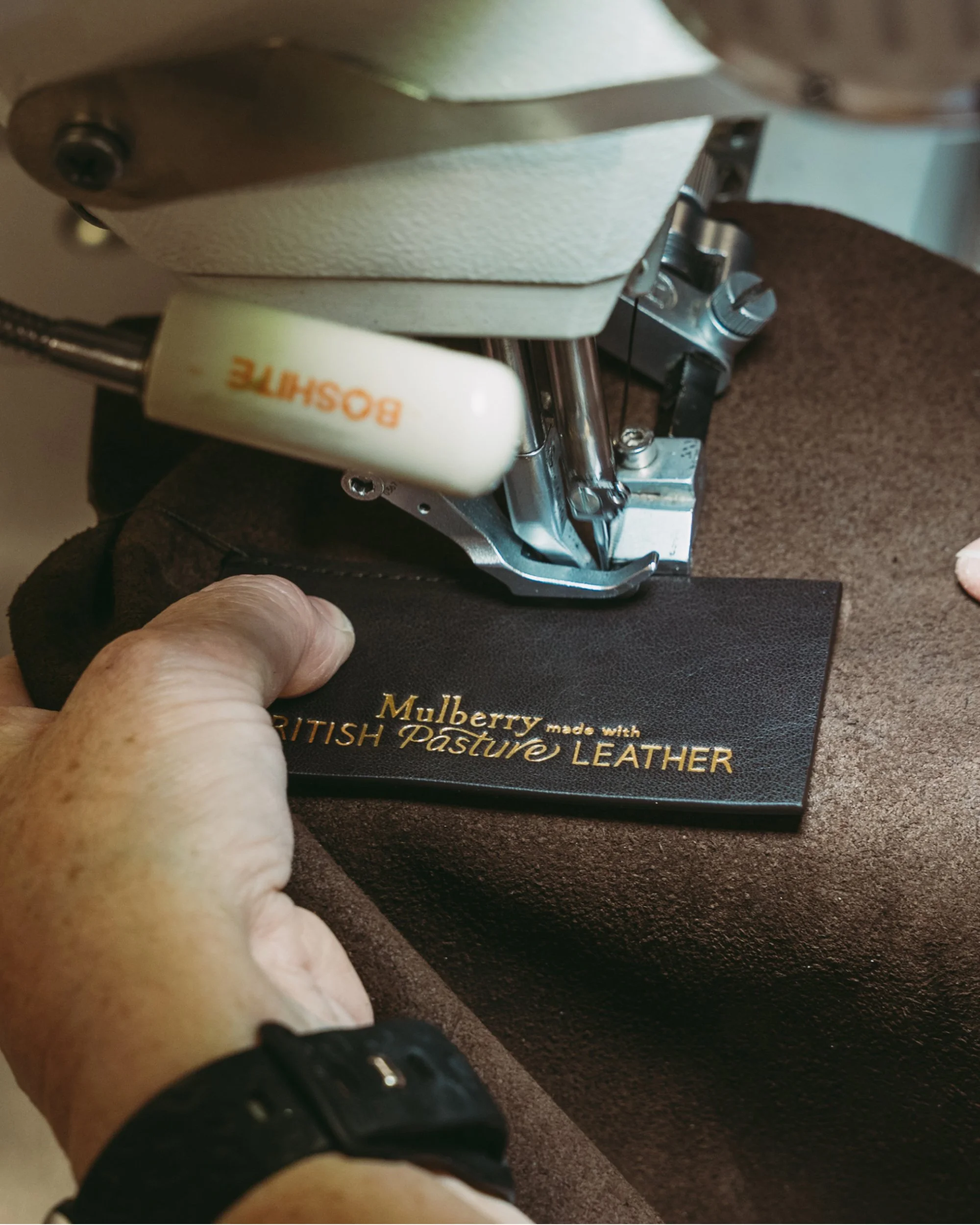 Close-up of a sewing machine needle and mechanism, with a hand holding a piece of black leather material beneath it, and a box labeled 'Mulberry made with British Pasture LEATHER' in the background.