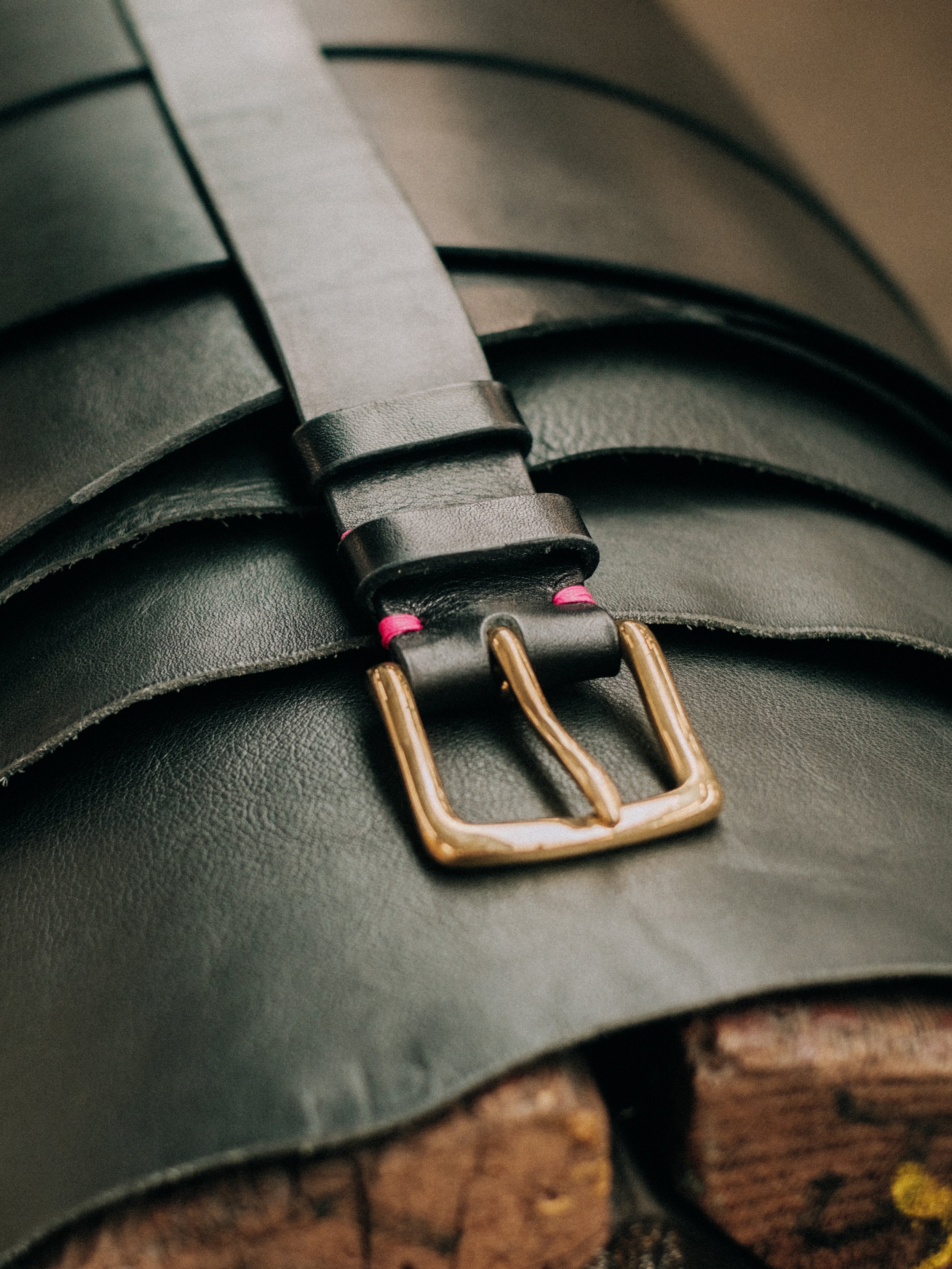 Close-up of a black leather belt with a gold buckle layered on top of other belts, with a wooden surface visible at the bottom.