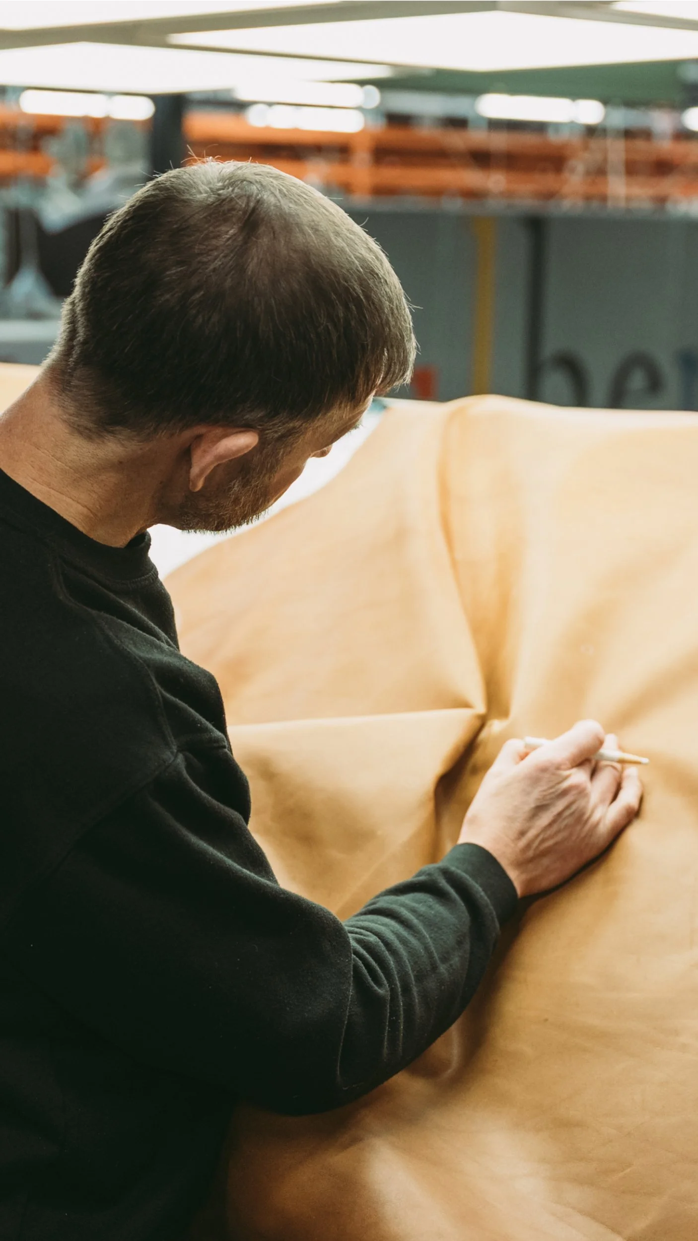 A man with brown hair and a beard working on a piece of light brown leather at a table in a workshop.