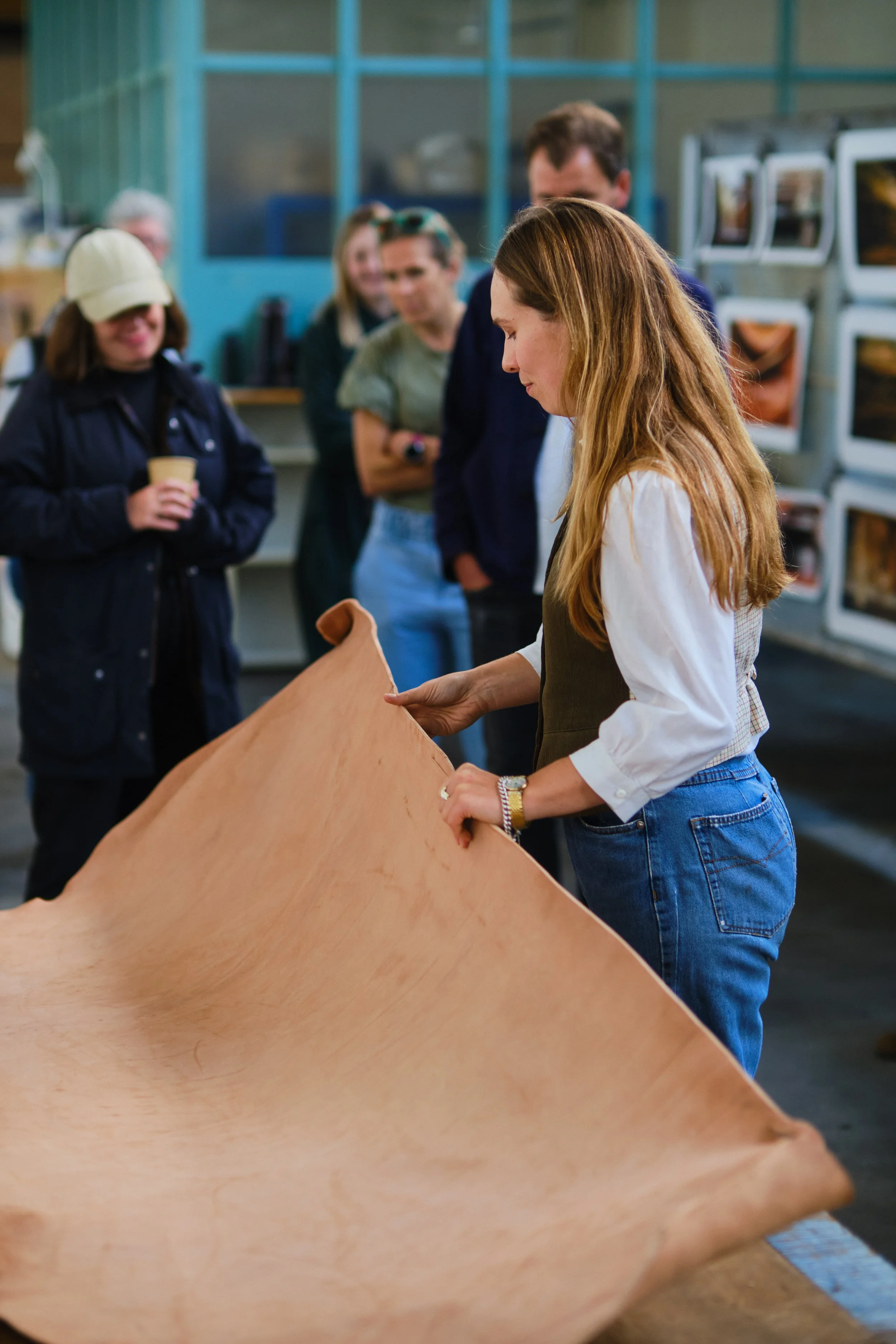A group of people gathered around a woman working on a large piece of leather in what appears to be a workshop or classroom.