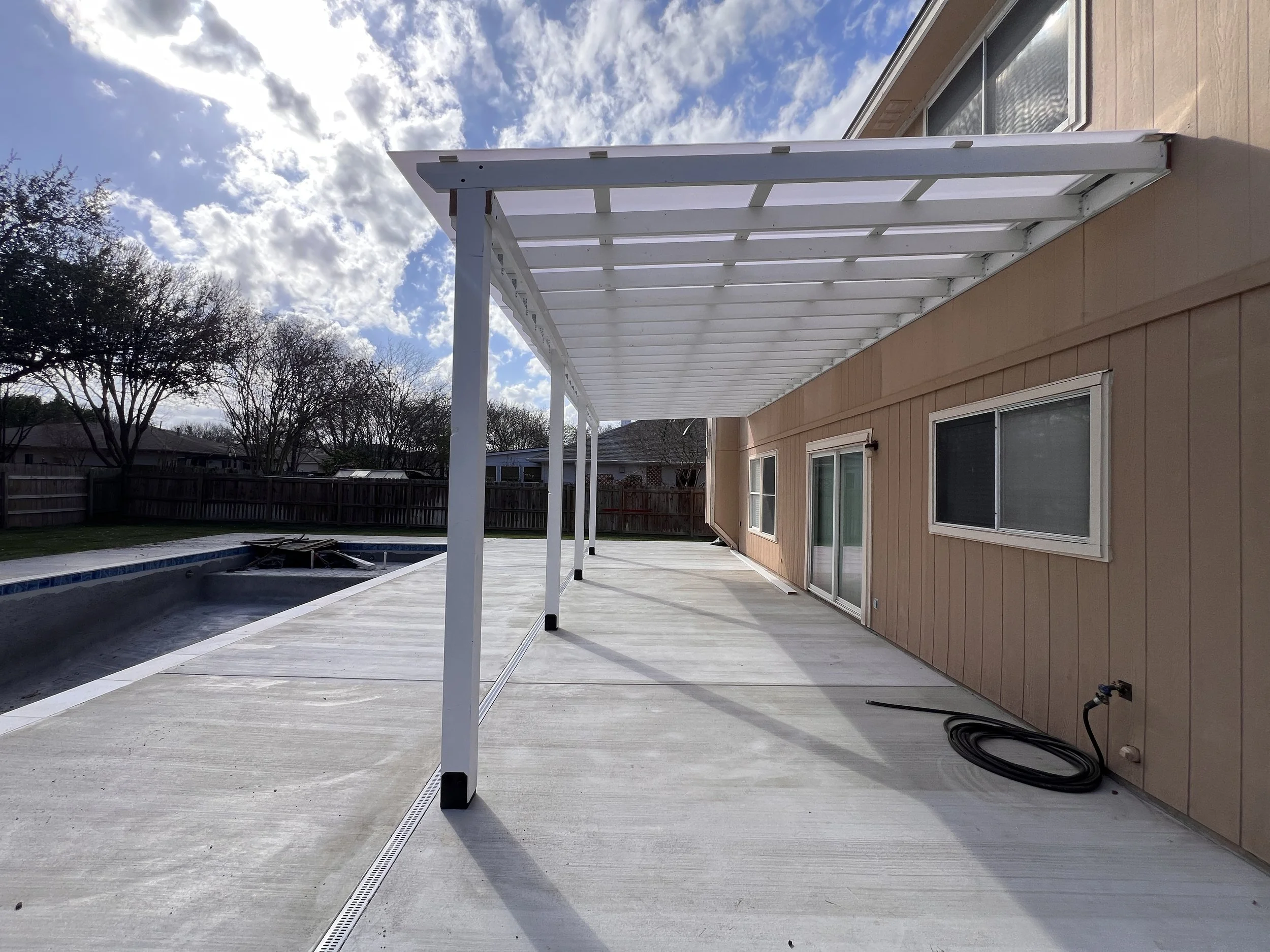New white patio cover with support beams over a concrete patio next to a beige house with sliding glass doors and two windows, a coiled garden hose on the ground, and a swimming pool with a liner underneath.