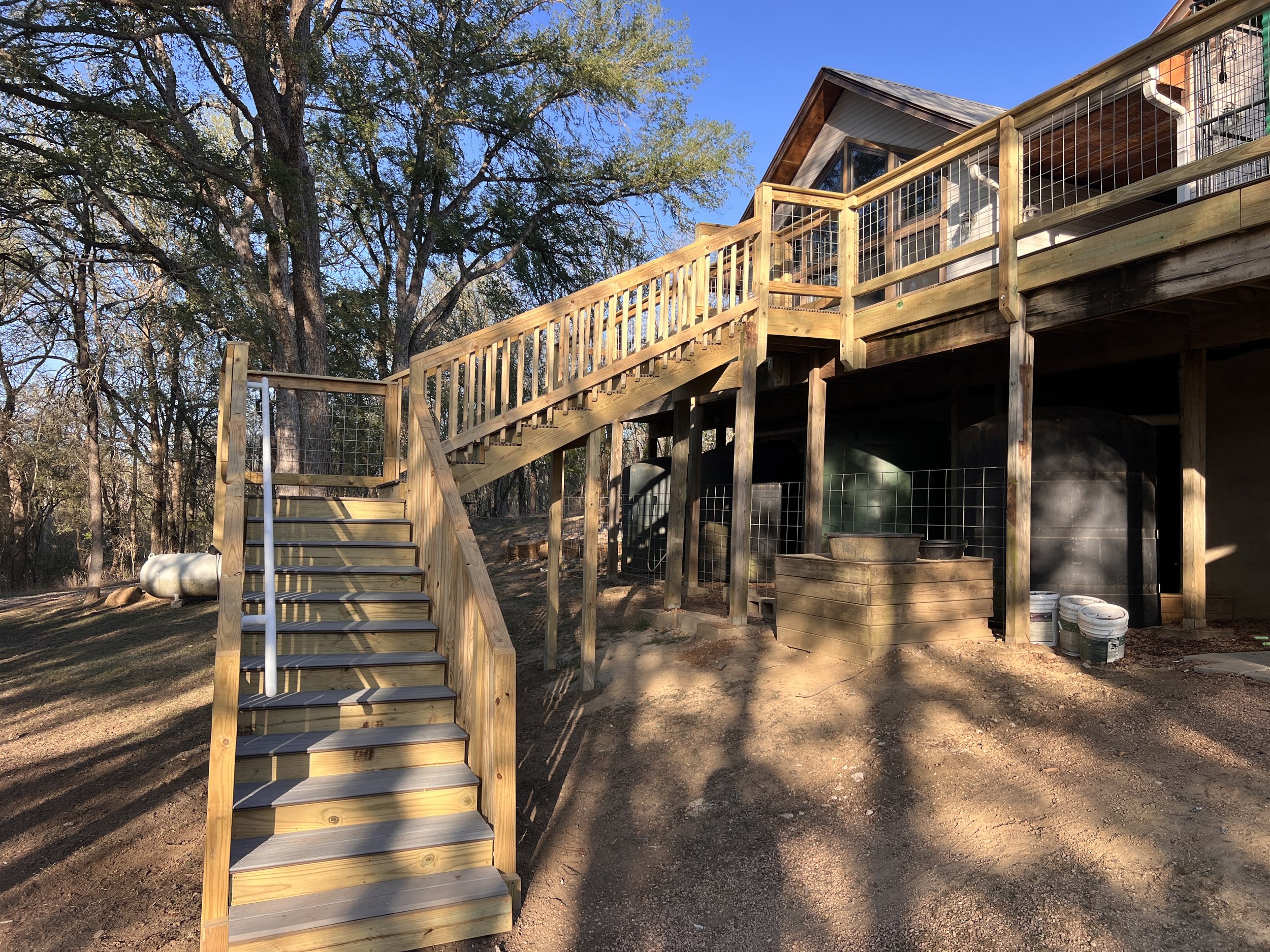 Wooden deck with stairs leading to a house, surrounded by trees on a sunny day.