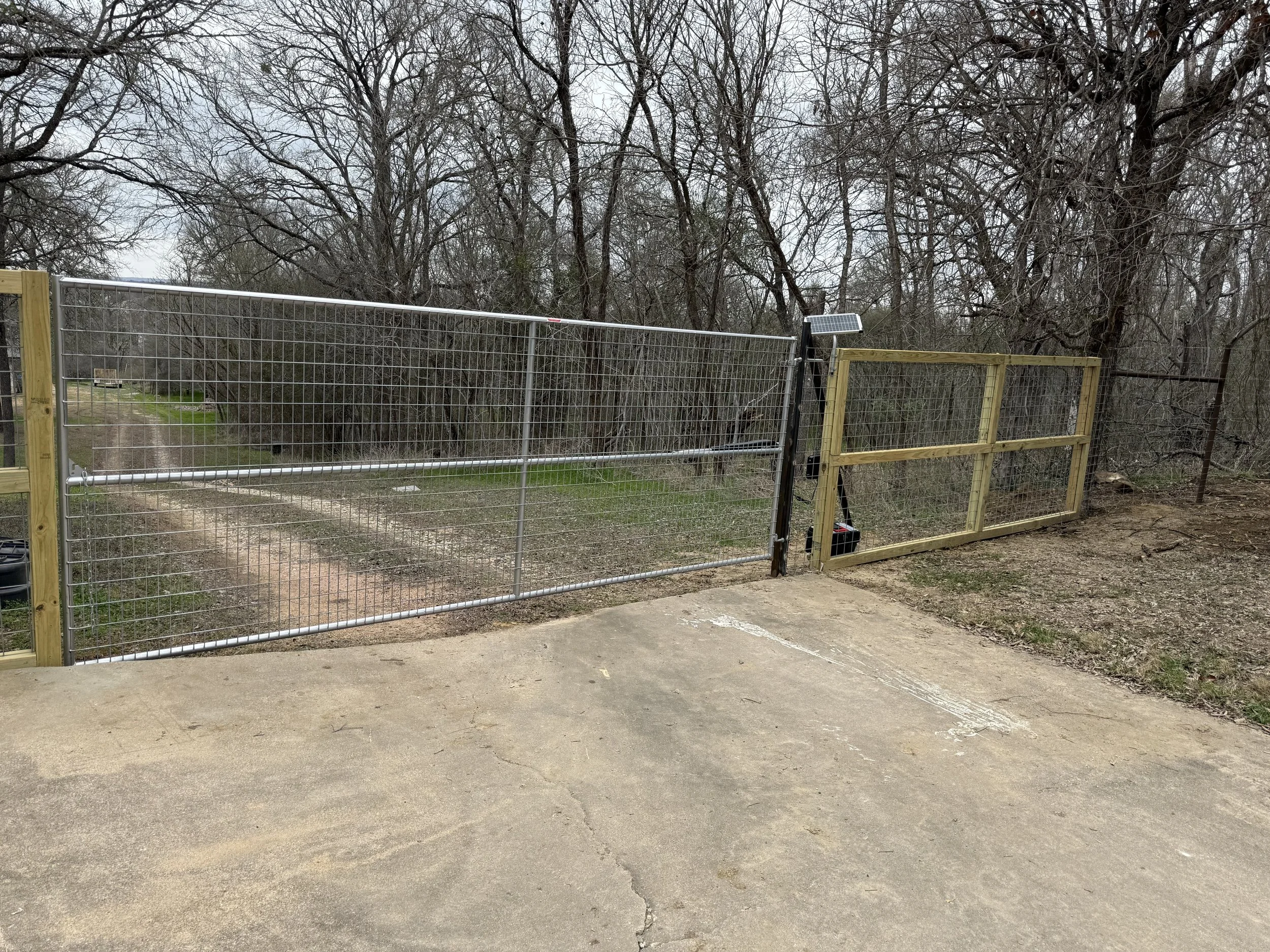 Wooden and metal gate with solar panel in front of a wooded area with leafless trees.
