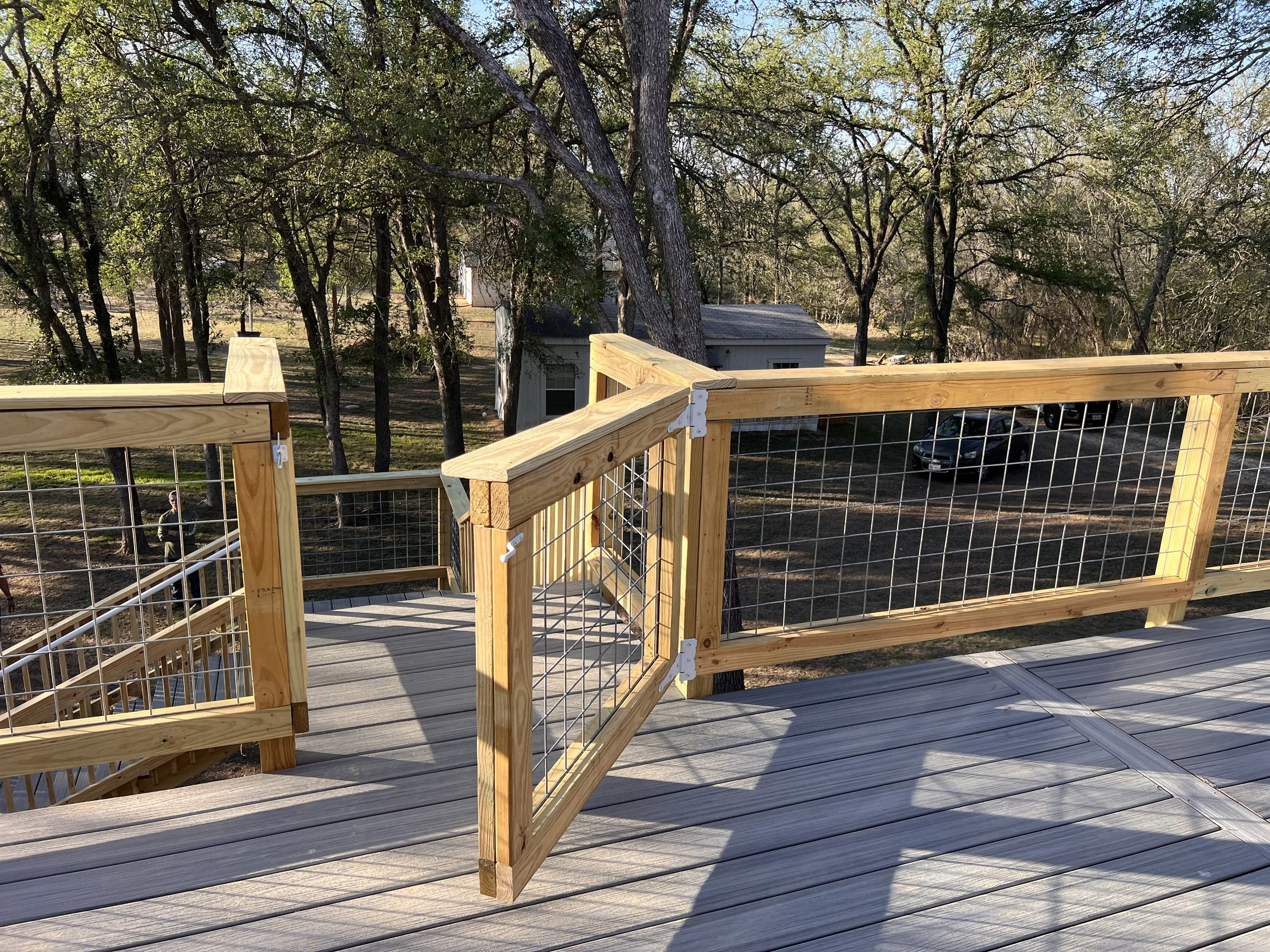 A newly built wooden deck with a railing overlooking a yard with trees, a small white shed, and parked cars. Two children are playing in the yard.
