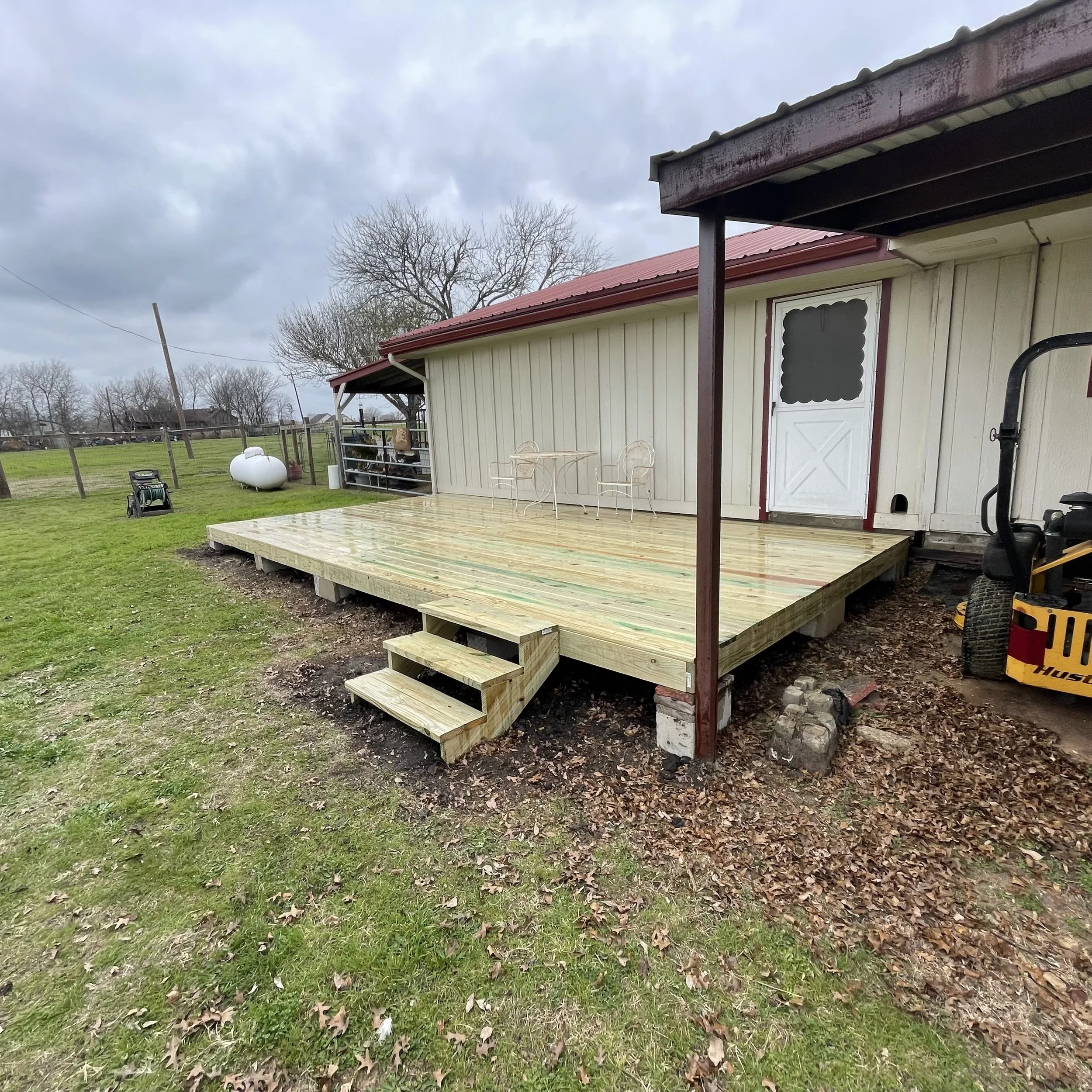 View of a wooden deck attached to a house with beige siding and a white door, in a backyard with green grass, leafless trees, and a cloudy sky.