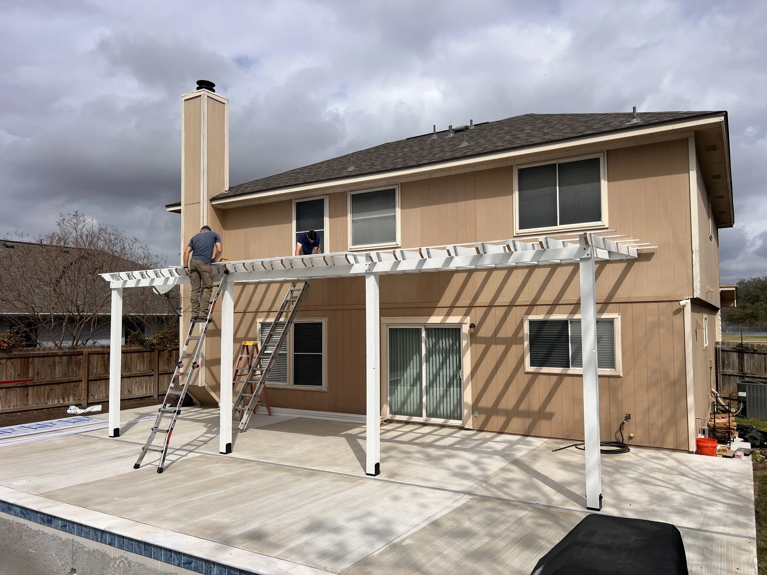 People assembling a white patio pergola on a backyard patio, with ladders and tools nearby, behind a beige house with a fenced yard.