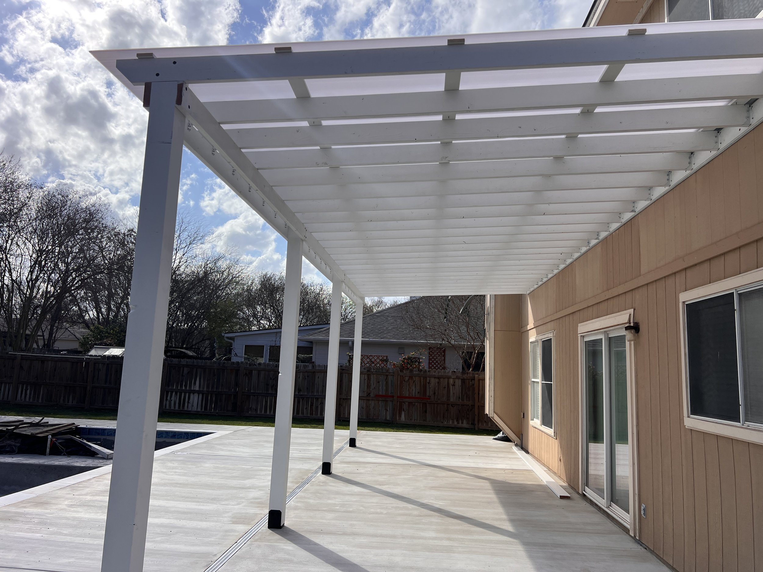Back patio with a newly built white pergola attached to a beige house, featuring a sliding glass door, windows, and a wooden fence in the background, under a partly cloudy sky.