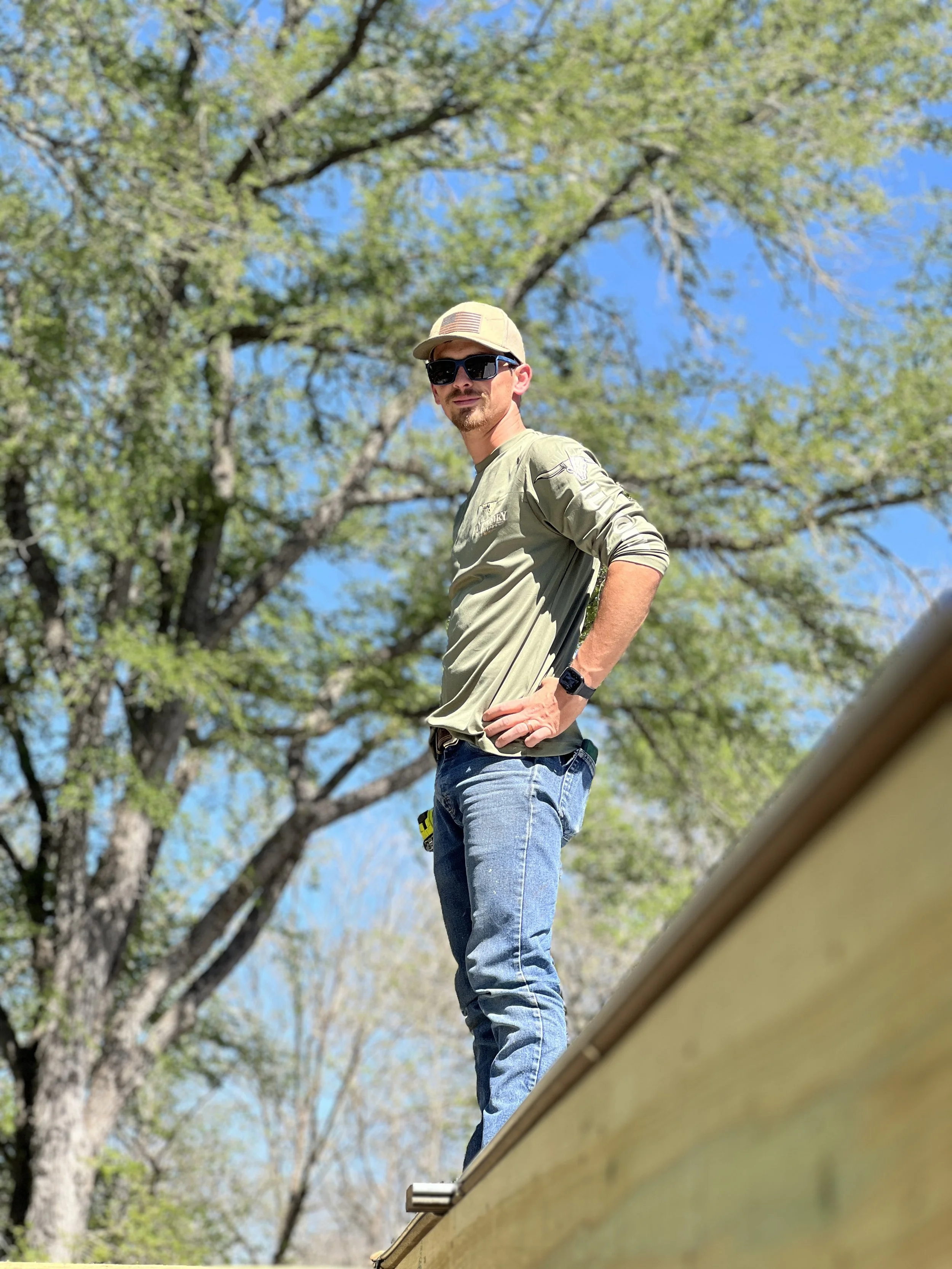 Man standing outdoors on a sunny day, wearing a baseball cap, sunglasses, a long-sleeve shirt, and jeans, with trees and blue sky in the background.