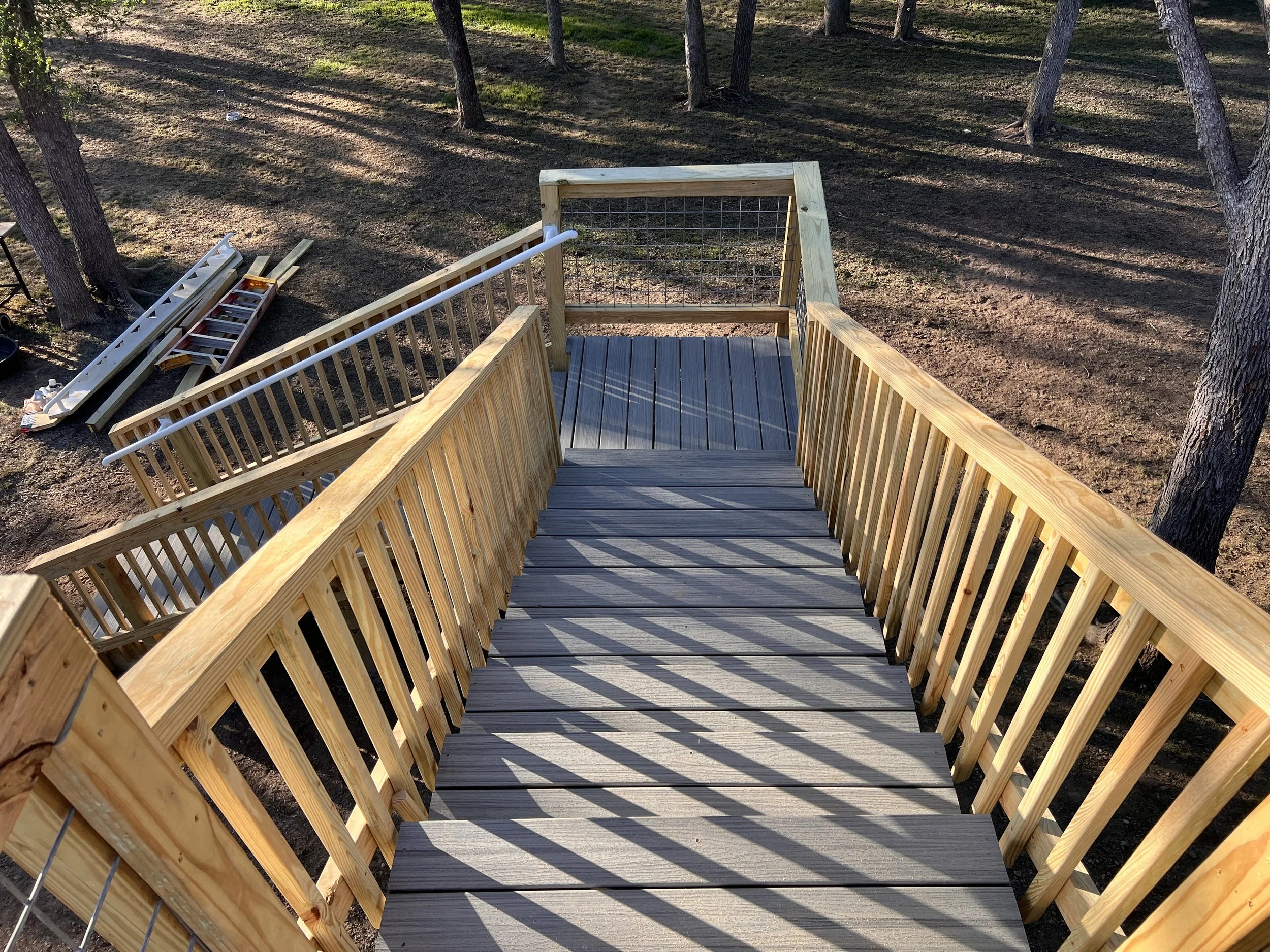 Wooden staircase outdoors with trees and shadows in the background, construction materials on the ground.