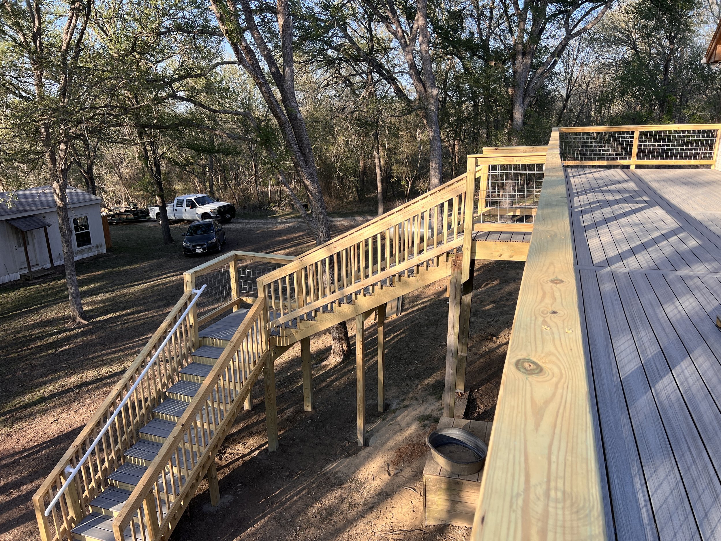 View from the top of a newly constructed wooden deck with stairs, overlooking a backyard with trees, a small shed, and parked cars.