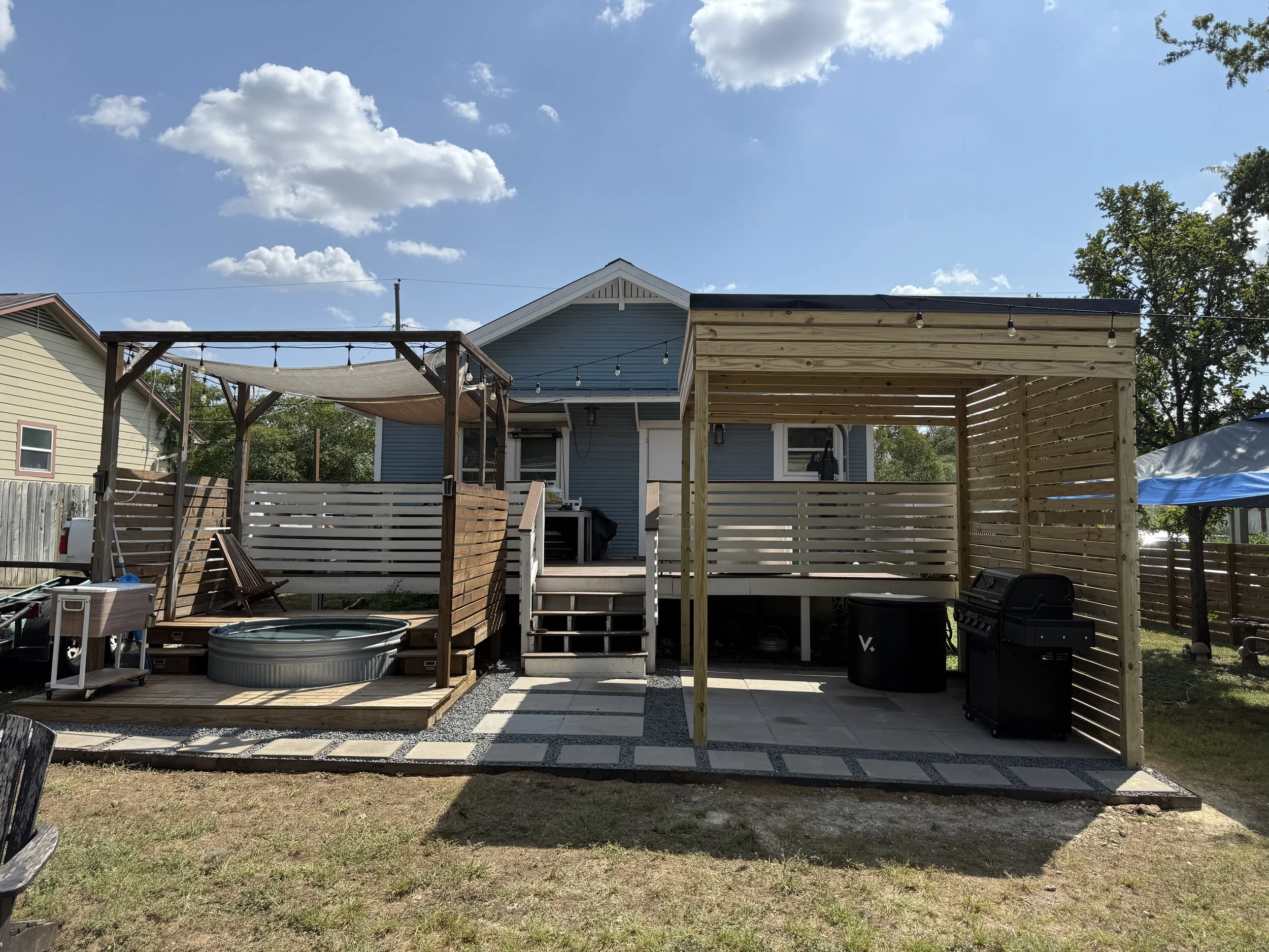 Backyard with a deck, two wooden pergolas, and a blue house. The sky is blue with some clouds.