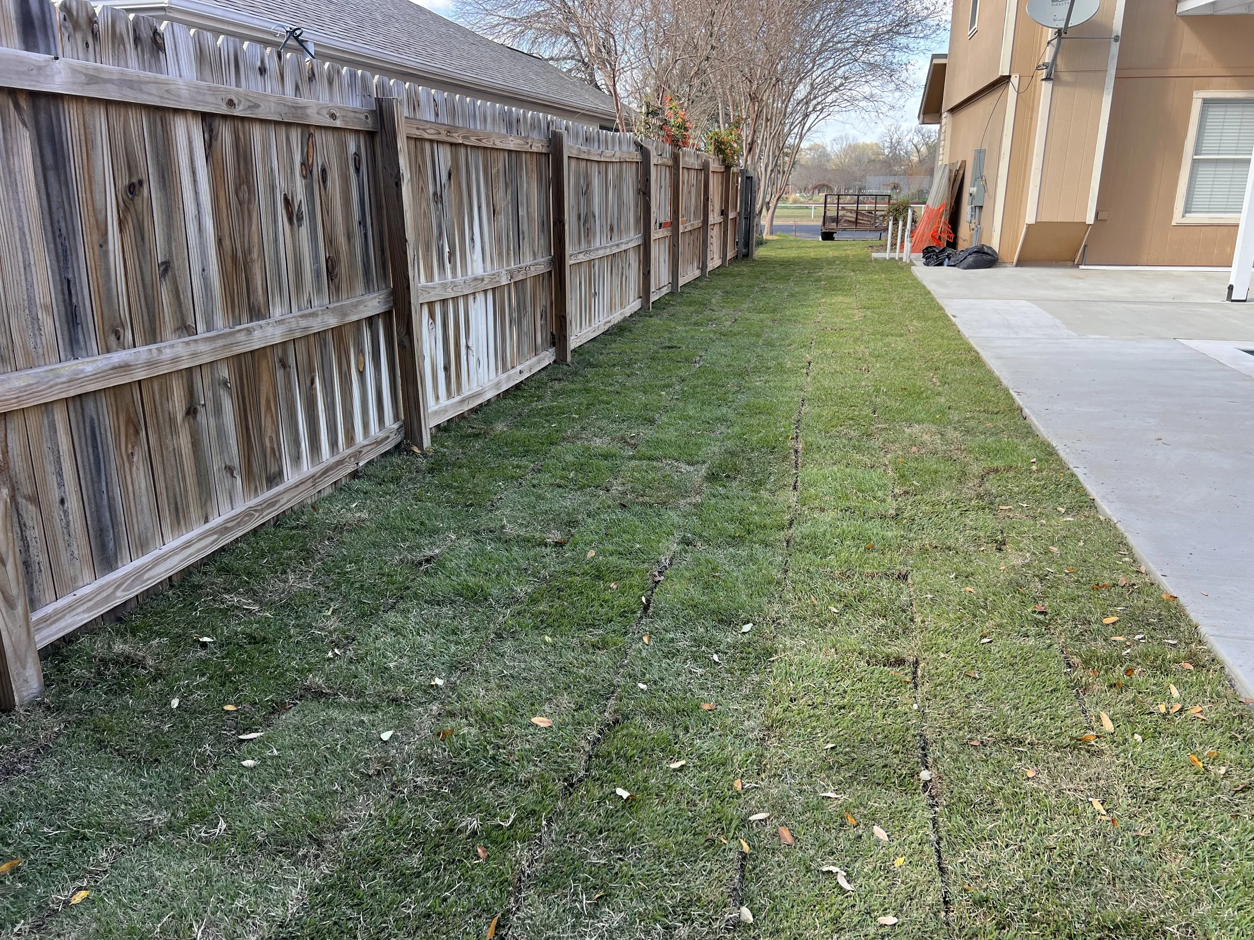 A backyard with a wooden fence, freshly laid sod, and a concrete patio area next to a house with brown siding. There are some black trash bags and orange safety fence on the side of the house.