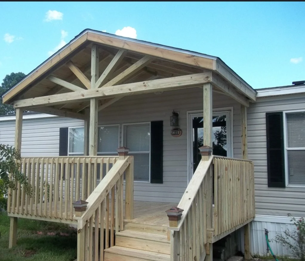 A house with a newly built wooden front porch and stairs, featuring a small roof over the porch supported by wooden posts, with windows and shutters visible on the house.