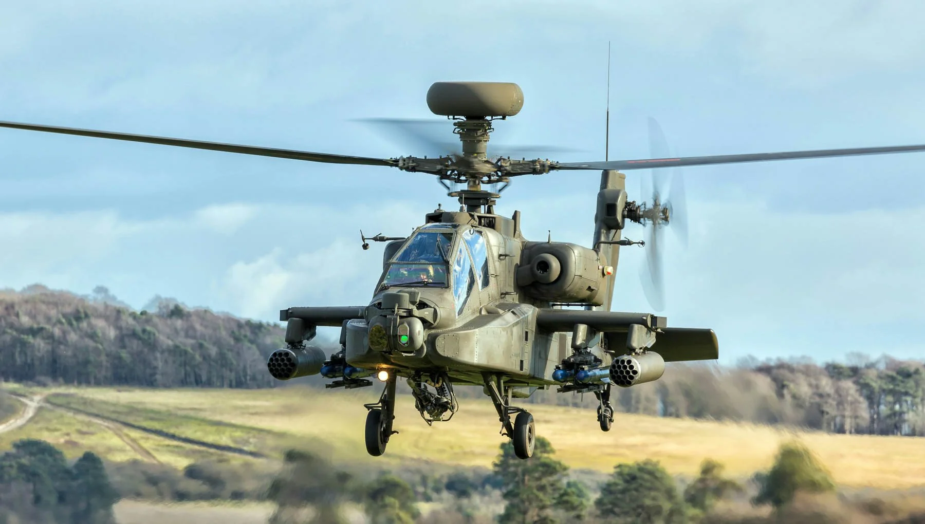A military attack helicopter flying low over a grassy landscape with trees and hills in the background.