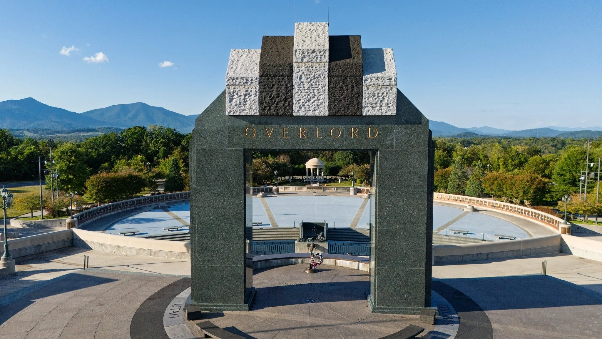 A monument with the word 'OVERLORD' in gold letters, with a large archway and steps leading to a circular plaza. In the background, a domed gazebo is visible amidst trees, with mountains and a blue sky.