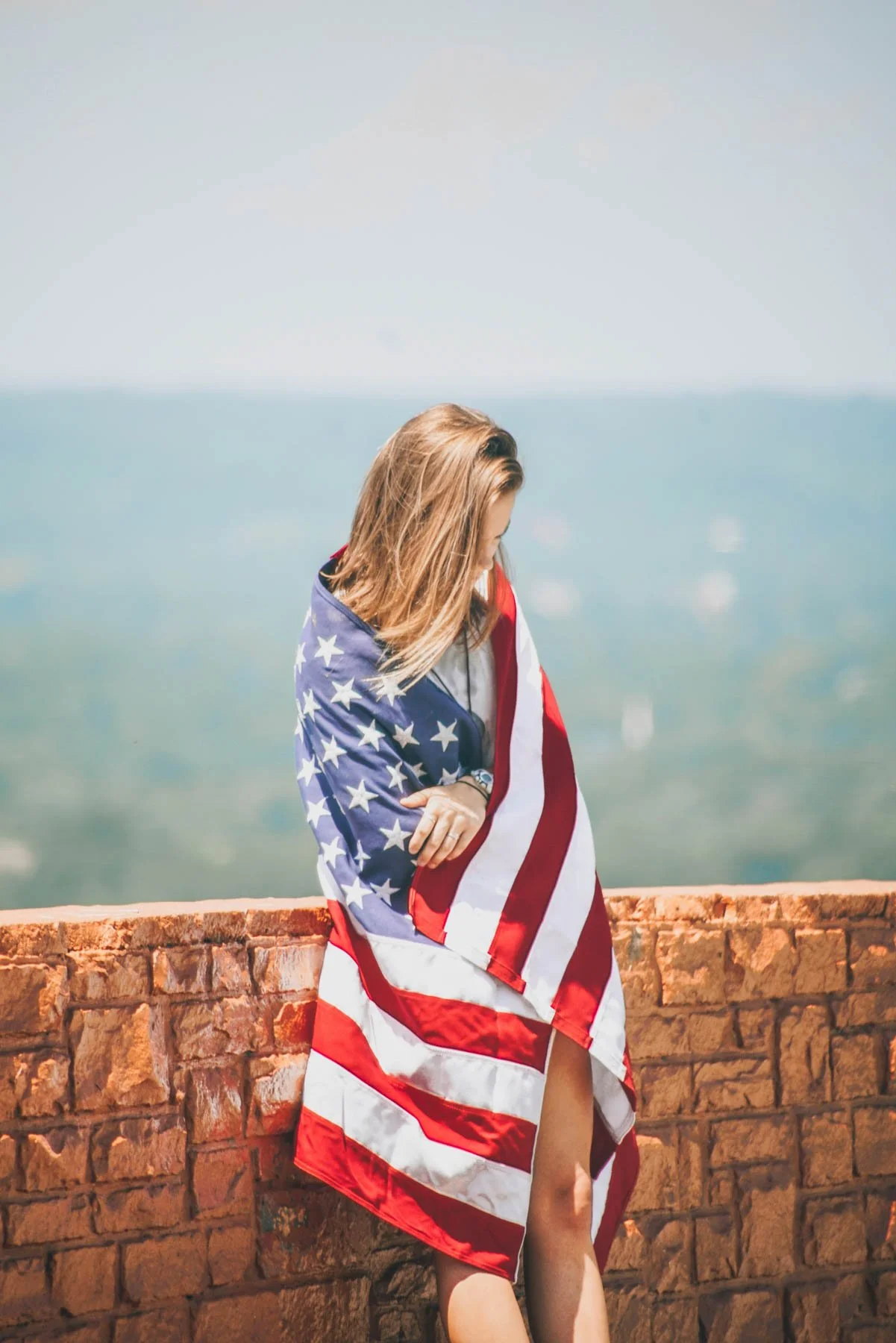 A woman standing on a brick wall with the American flag draped over her shoulders, looking down with her hair covering her face, in front of a blurry background of blue sky and water.