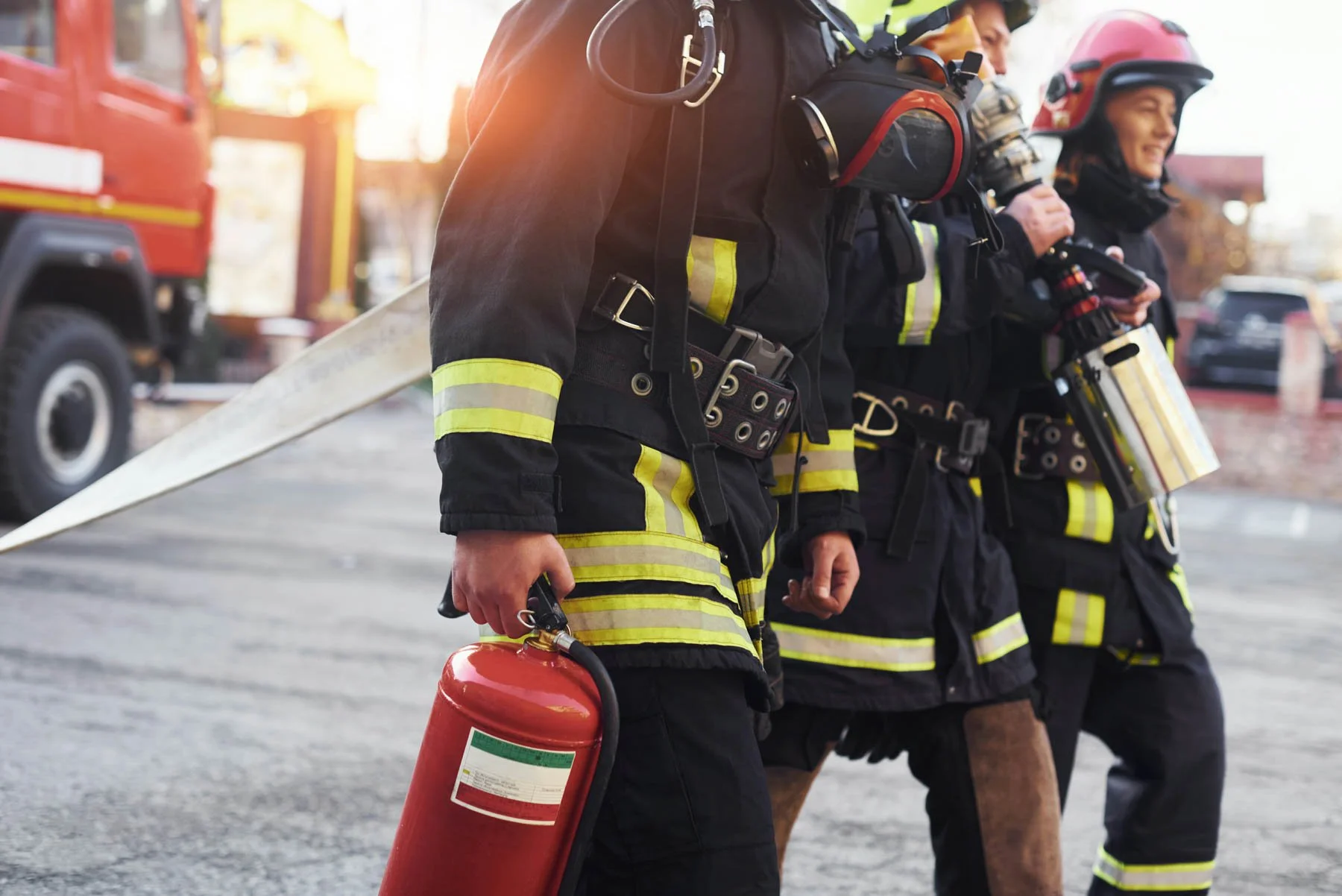 Firefighters walking outdoors, with one carrying a fire extinguisher, all wearing protective gear and helmets.