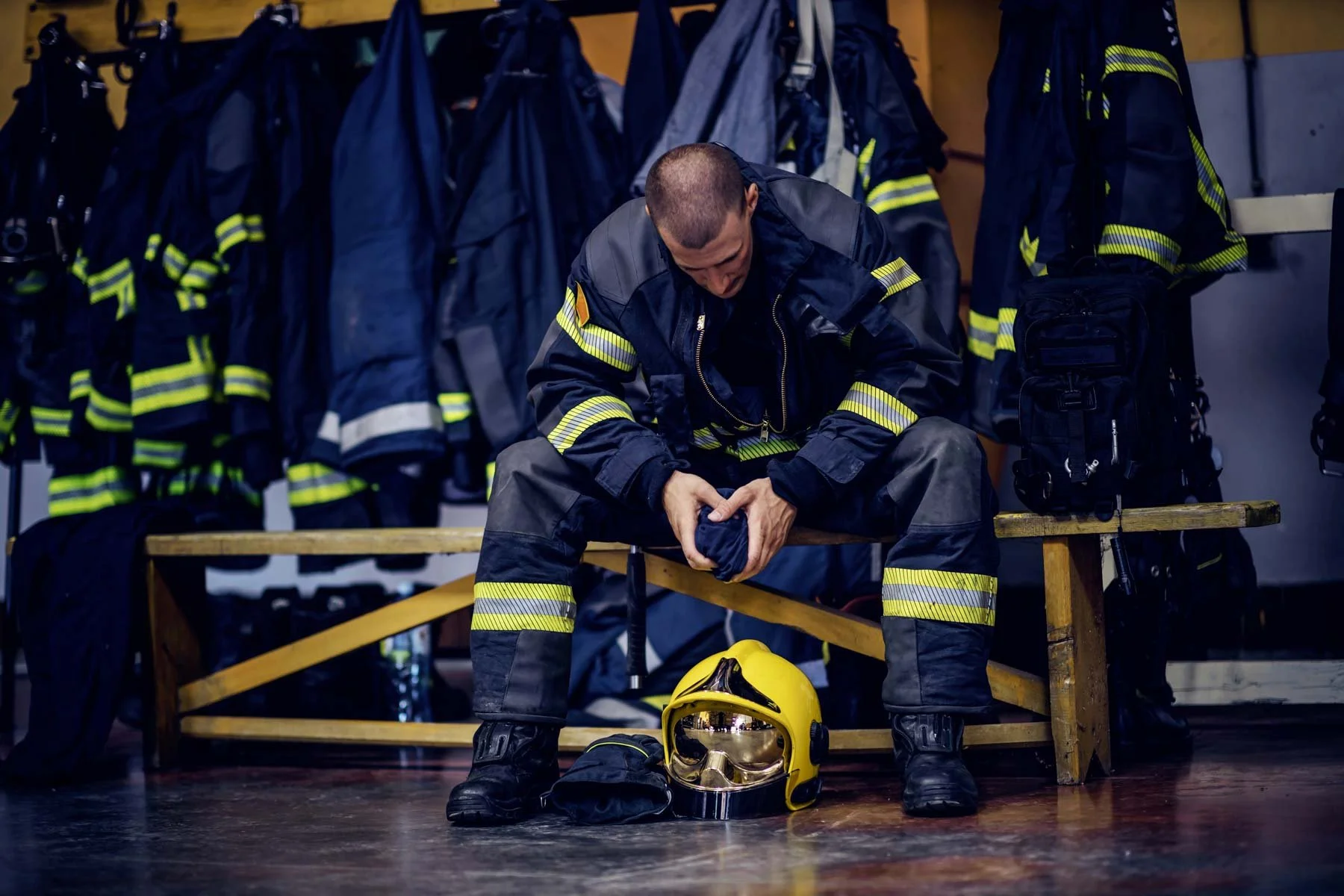 Firefighter in uniform putting on black gloves while sitting on a wooden bench in firehouse, with yellow helmet and face shield on the floor, and additional gear hanging on the wall behind.