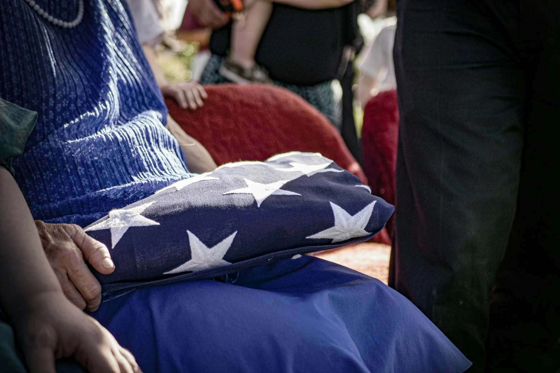 Person holding a folded American flag on their lap, with other people and chairs in the background.