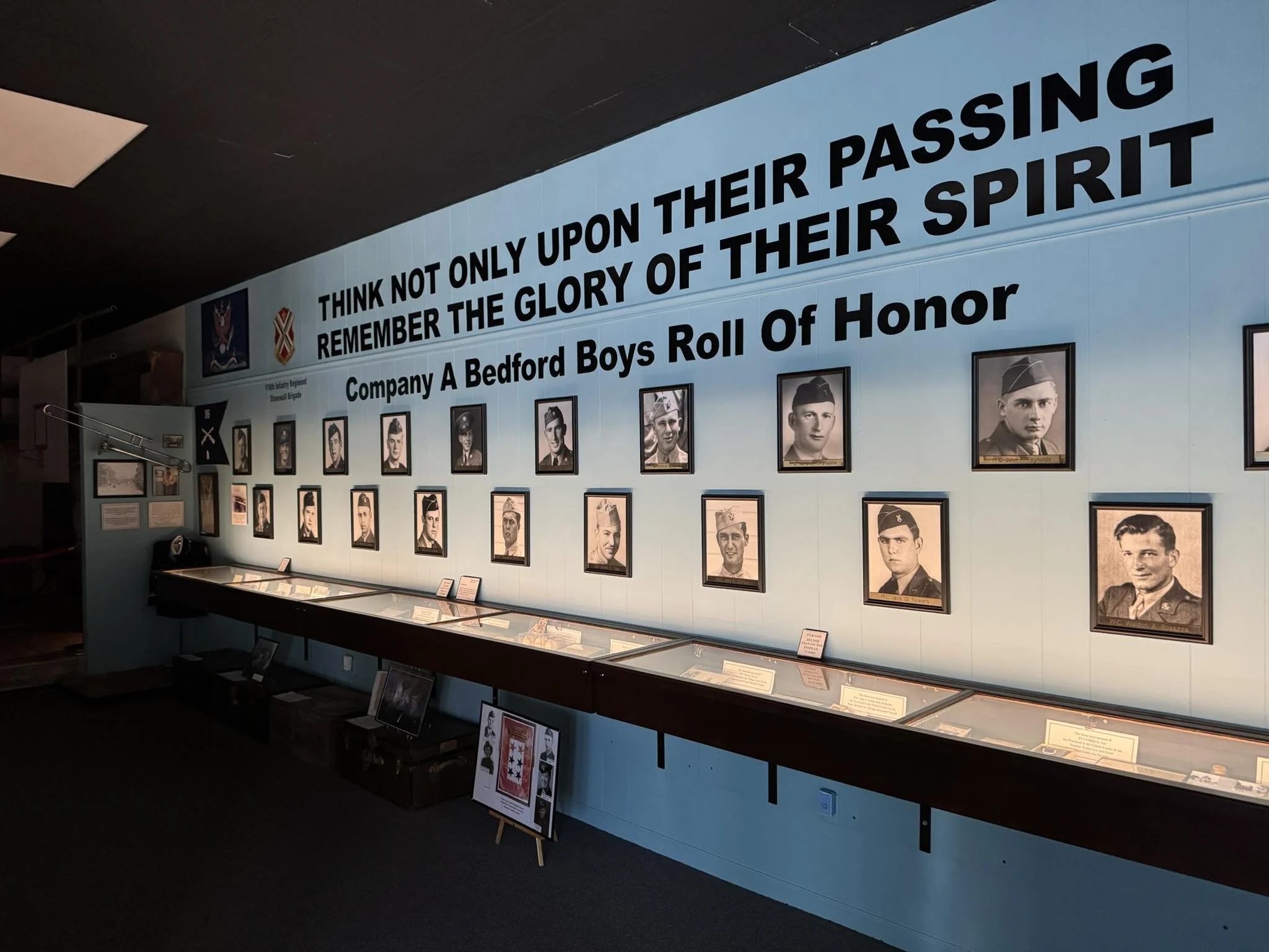 Inside a museum exhibit with portraits of soldiers, a large blue wall with a quote about their passing, and a display case with artifacts related to Company A Bedford Boys.