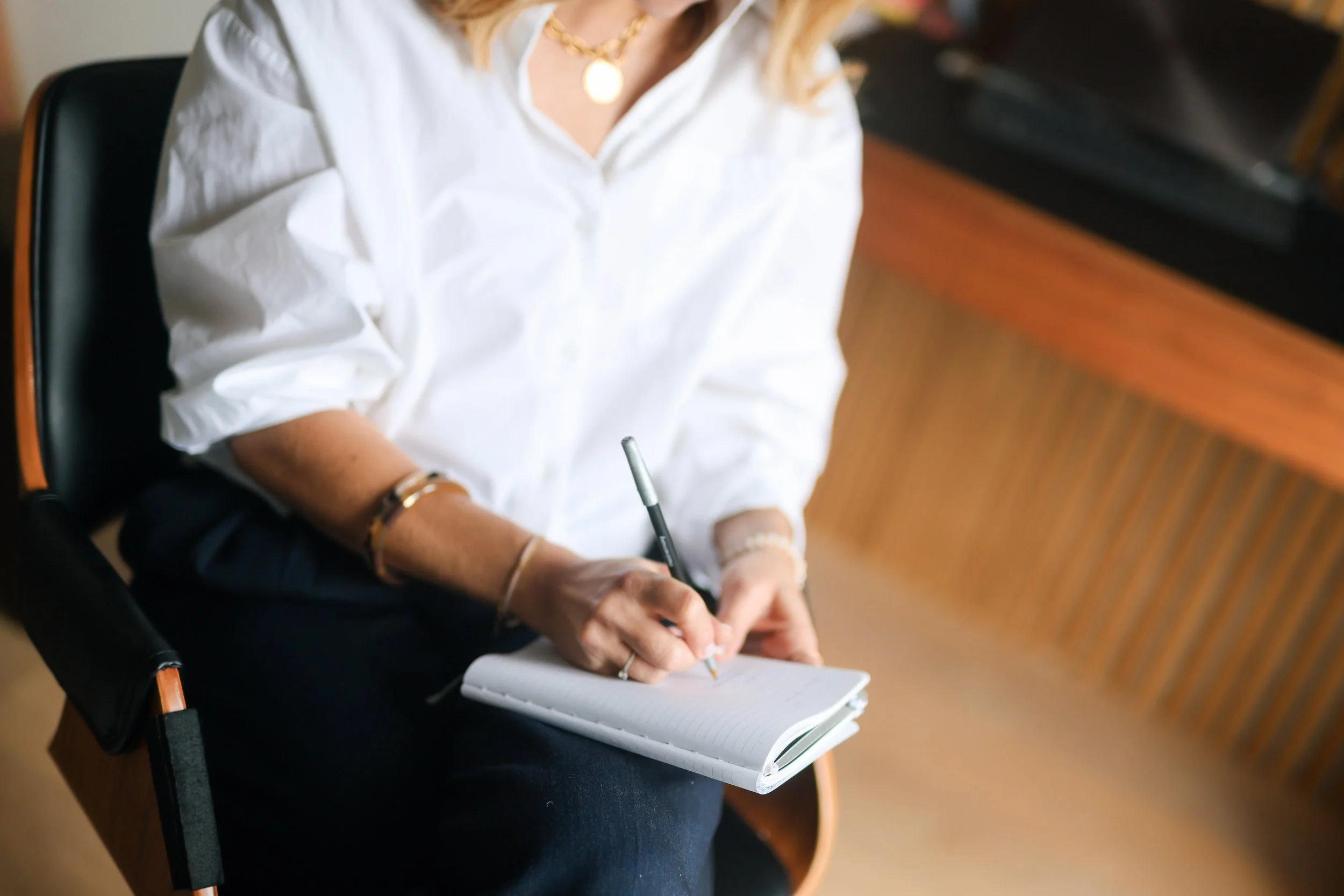 A woman sitting on a chair, wearing a white blouse, holding a pen, and writing in a notebook.