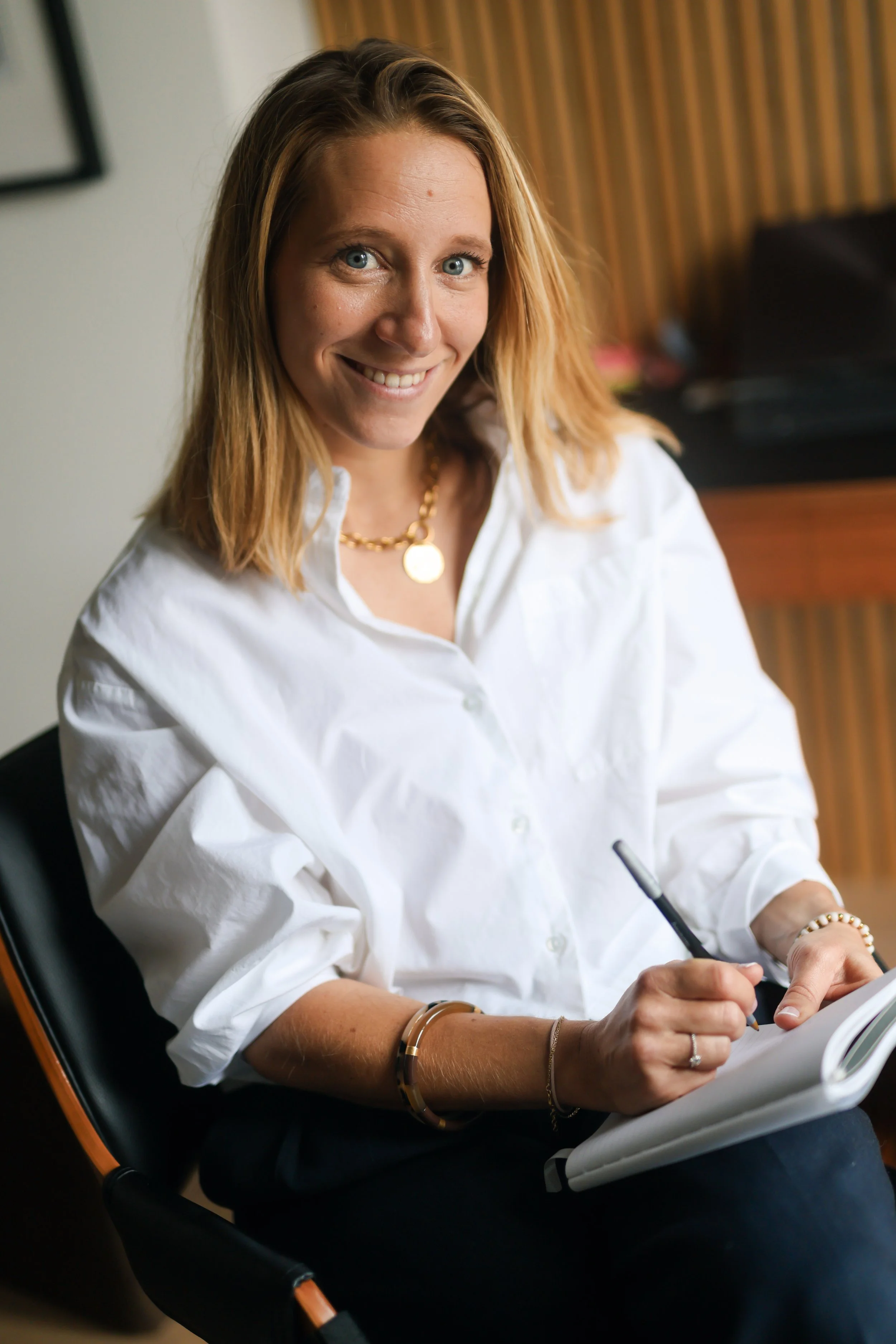 A woman with blonde hair and blue eyes smiling while sitting in a black and orange chair, wearing a white shirt, gold jewelry, and holding a pen and notebook.