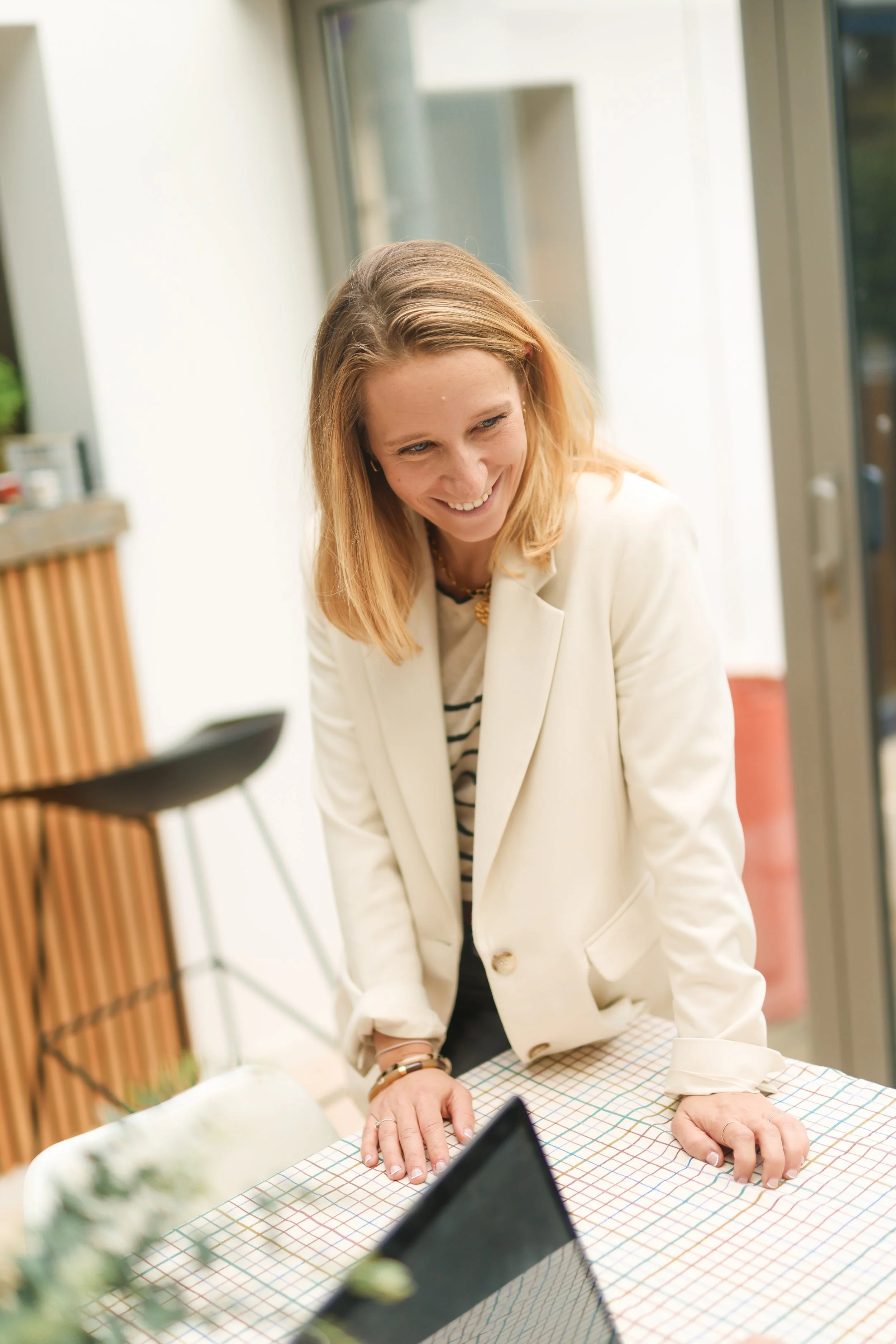 A woman with blonde hair, wearing a white blazer and striped shirt, smiling while leaning on a checked table in a bright room.