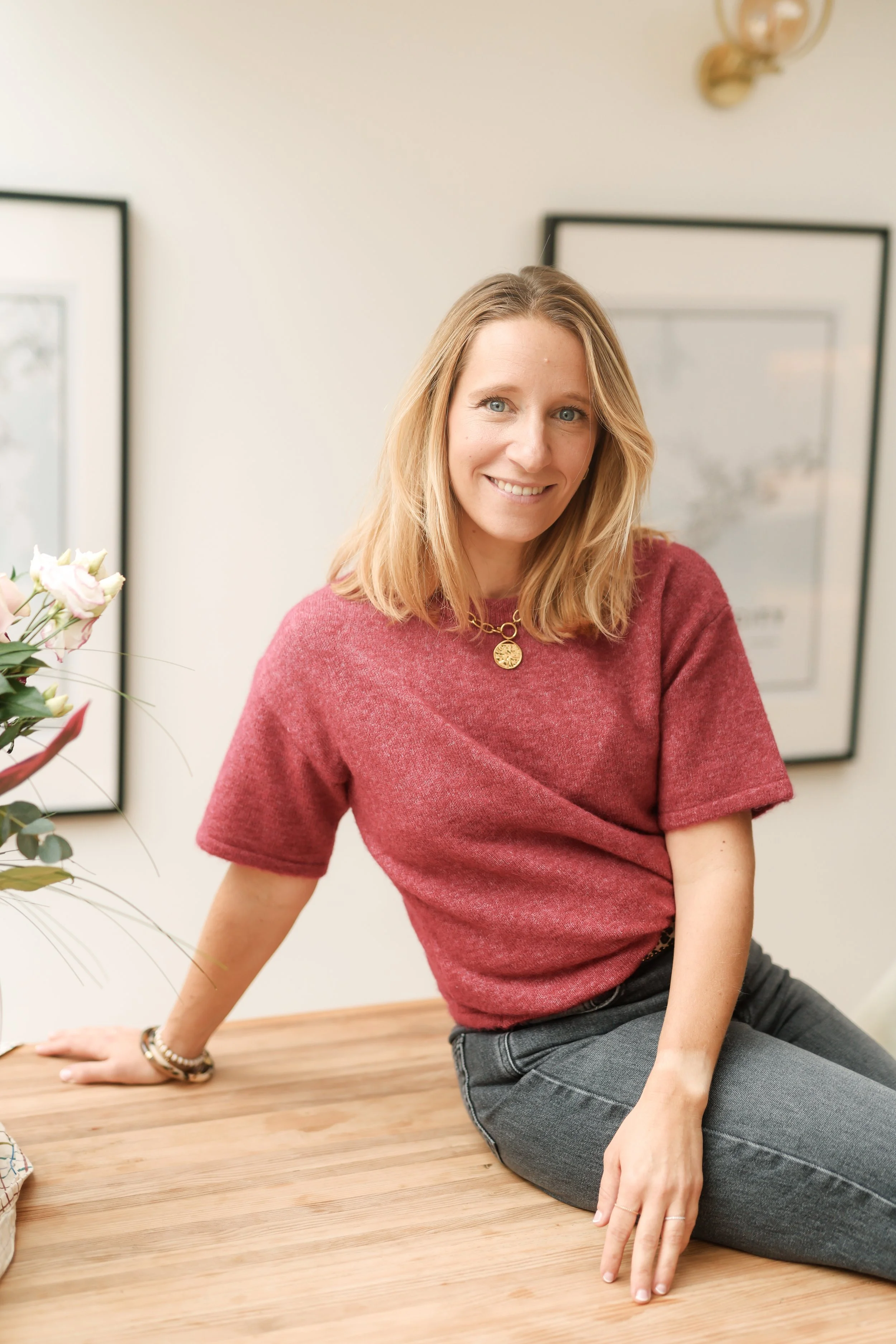 A woman with blonde hair smiling, sitting on a wooden table in a room with framed pictures on the wall and a flower arrangement nearby.