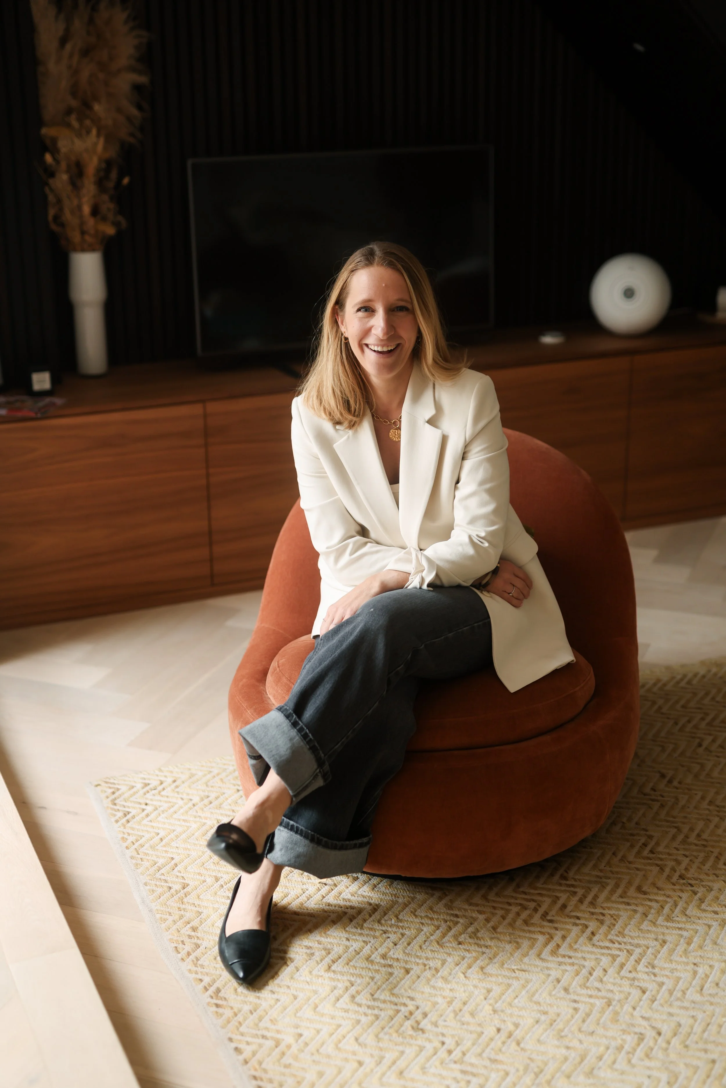 A woman sitting on a rounded orange armchair in a stylish living room, smiling at the camera. She is wearing a white blazer, dark jeans, and black shoes. Behind her, there's a wooden cabinet with decorative objects, including a white vase with dried 