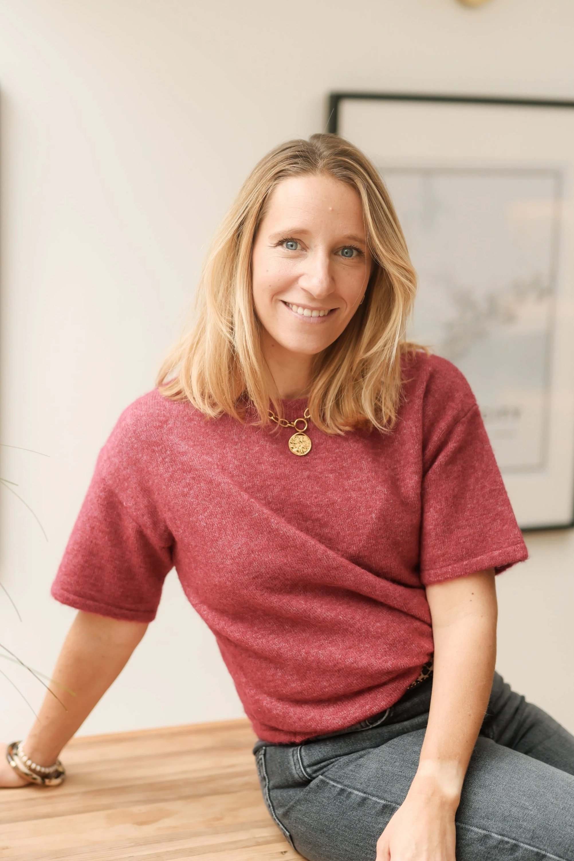 A smiling woman with shoulder-length blonde hair, wearing a red short-sleeved top and gray jeans, sitting on a wooden surface with her left arm resting on it, in a room with a framed picture behind her.