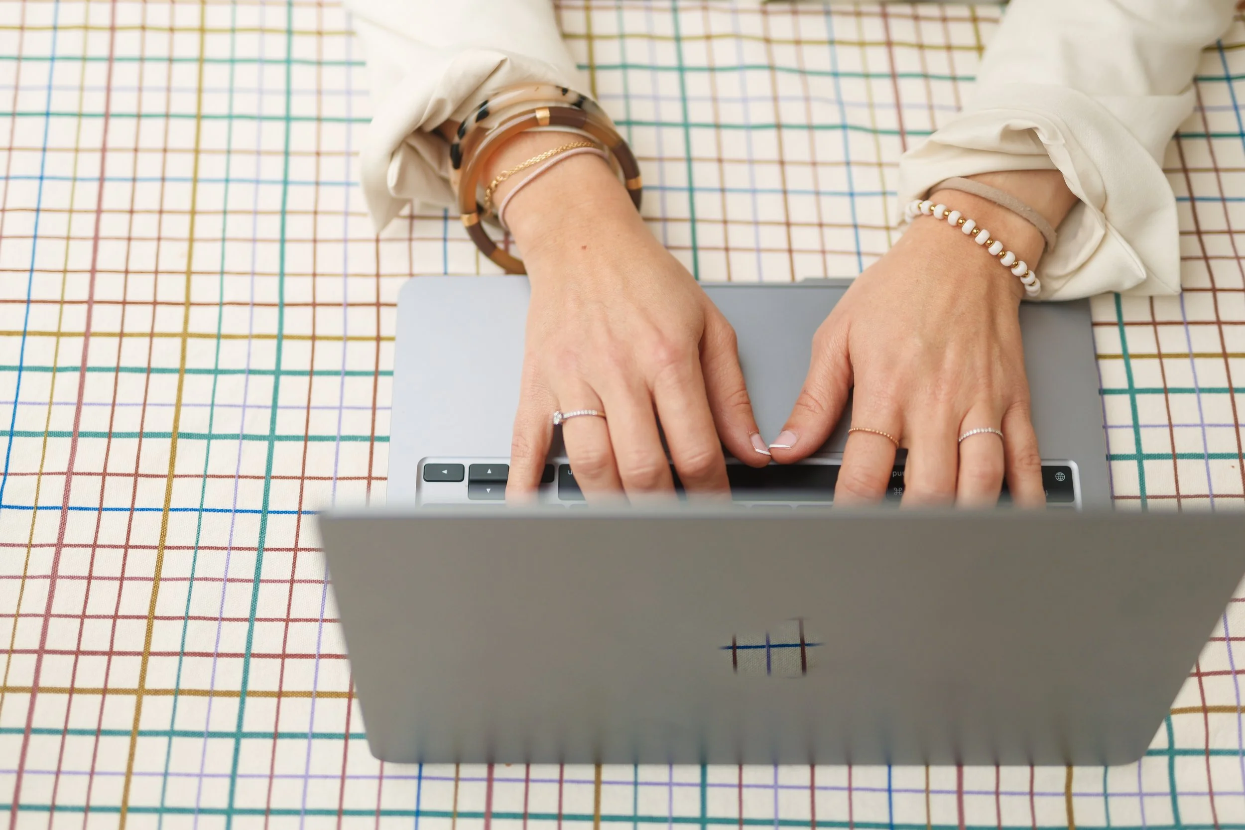 Person typing on a laptop with several bracelets on their wrists, sitting at a table with a colorful grid pattern.