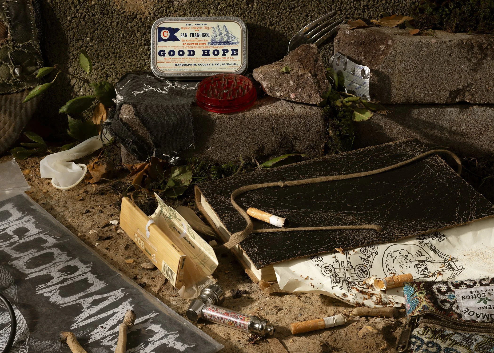 A cluttered surface with various objects including a vintage San Francisco ship souvenir box, a small red plastic item with spikes, a broken brick, plants, scattered tubes, a black cloth, and other miscellaneous items.