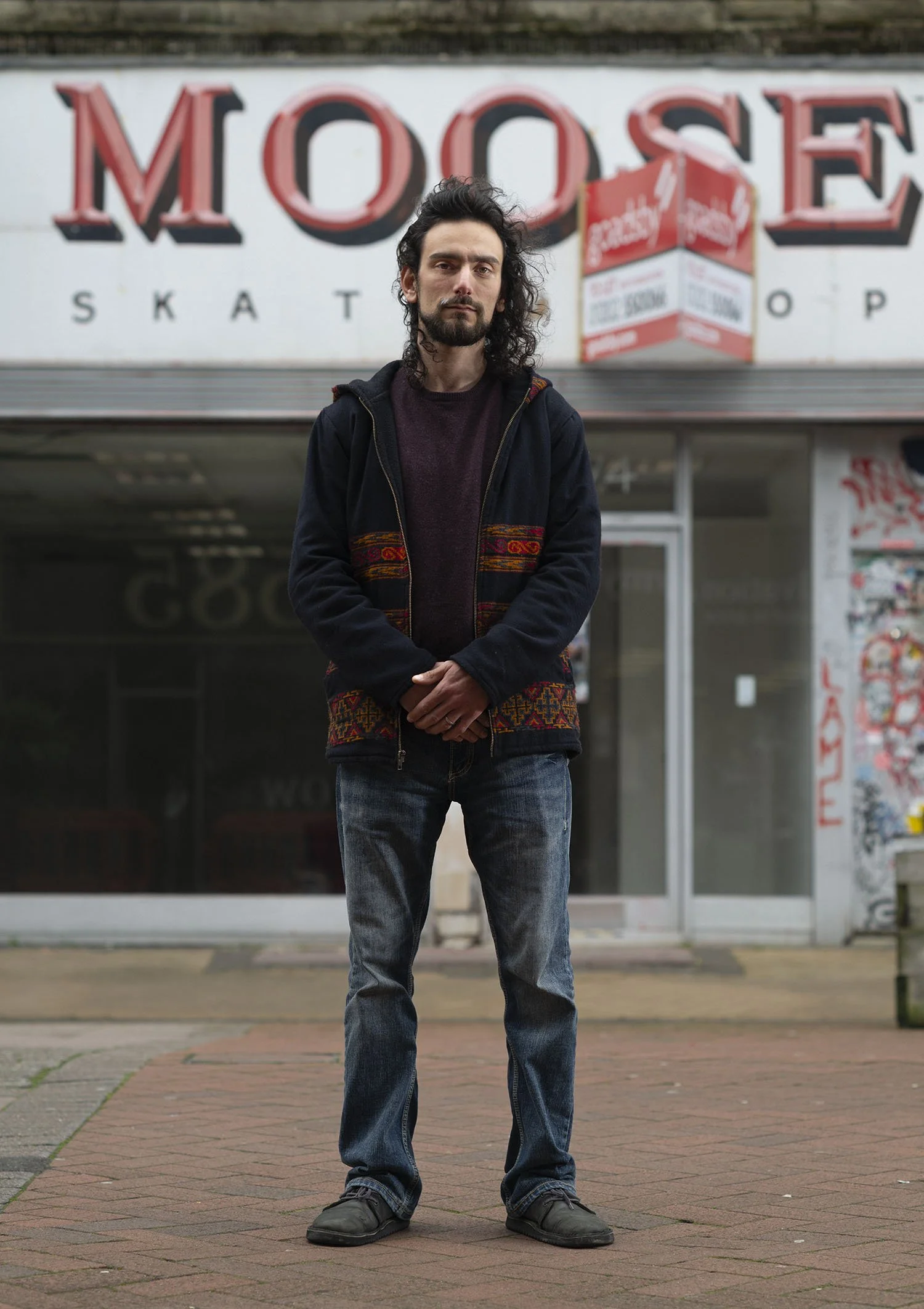 A man with dark, curly hair and a beard stands on a sidewalk outside a store with a large sign reading 'MOOSE SKATE'. He is wearing a dark jacket with a colorful pattern on the bottom, a maroon sweater, and jeans. He has a serious expression and is s