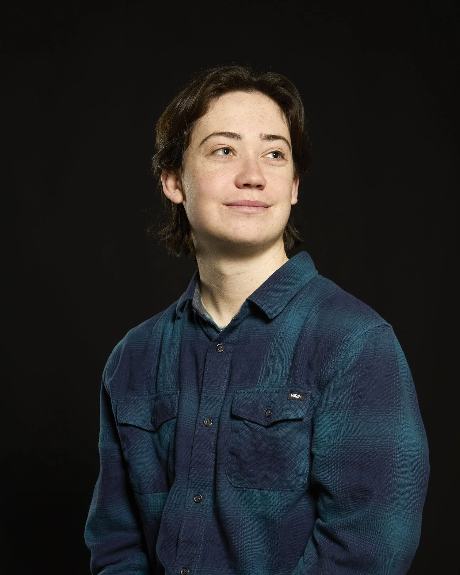 Nick Shorten Headshot with brown, shoulder-length hair, light skin with freckles, wearing a blue flannel shirt, looking to the side against a black background.