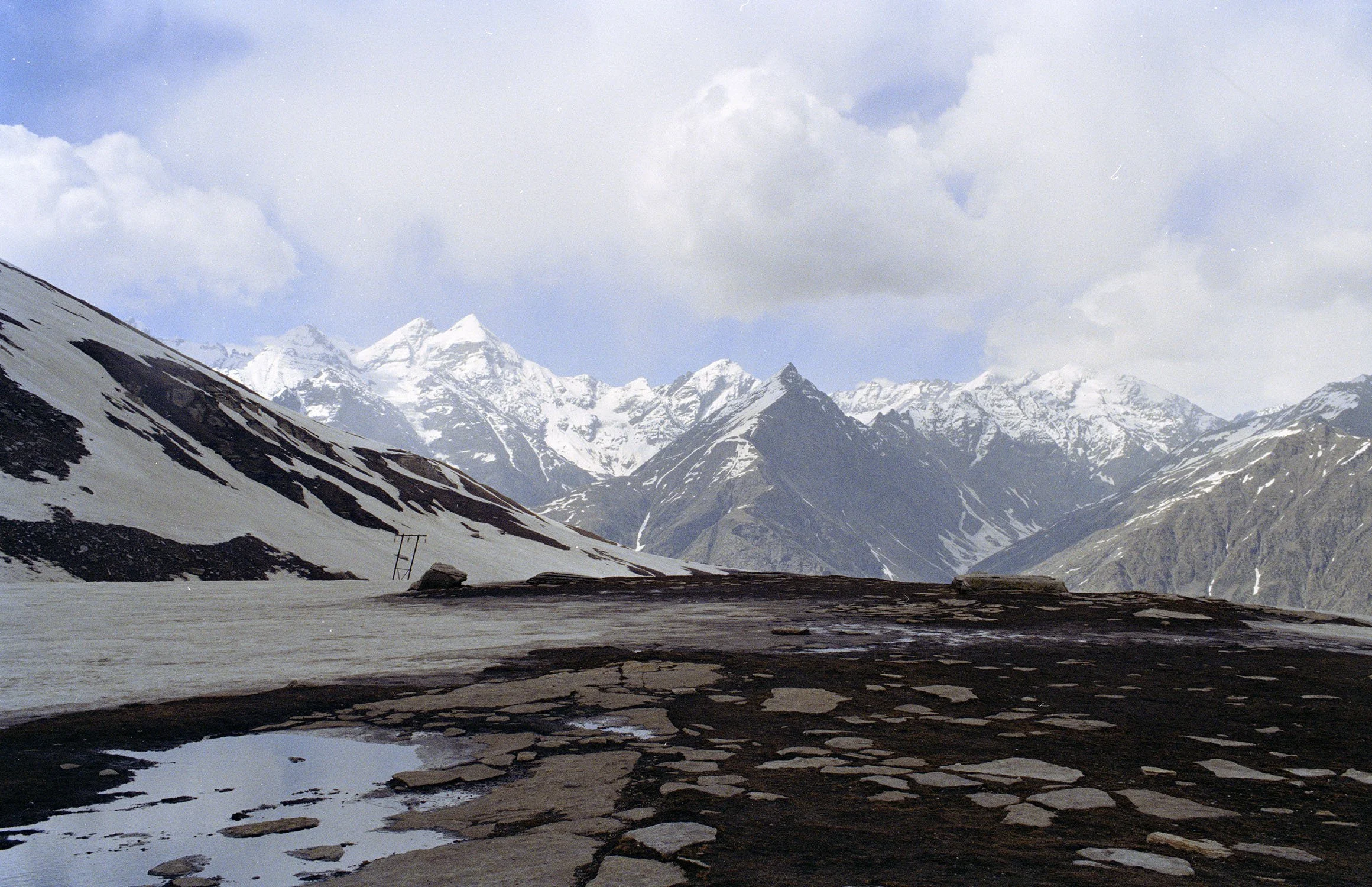 Himalayas under a cloudy sky with patches of snow and rock on the slopes and a frozen body of water in the foreground.
