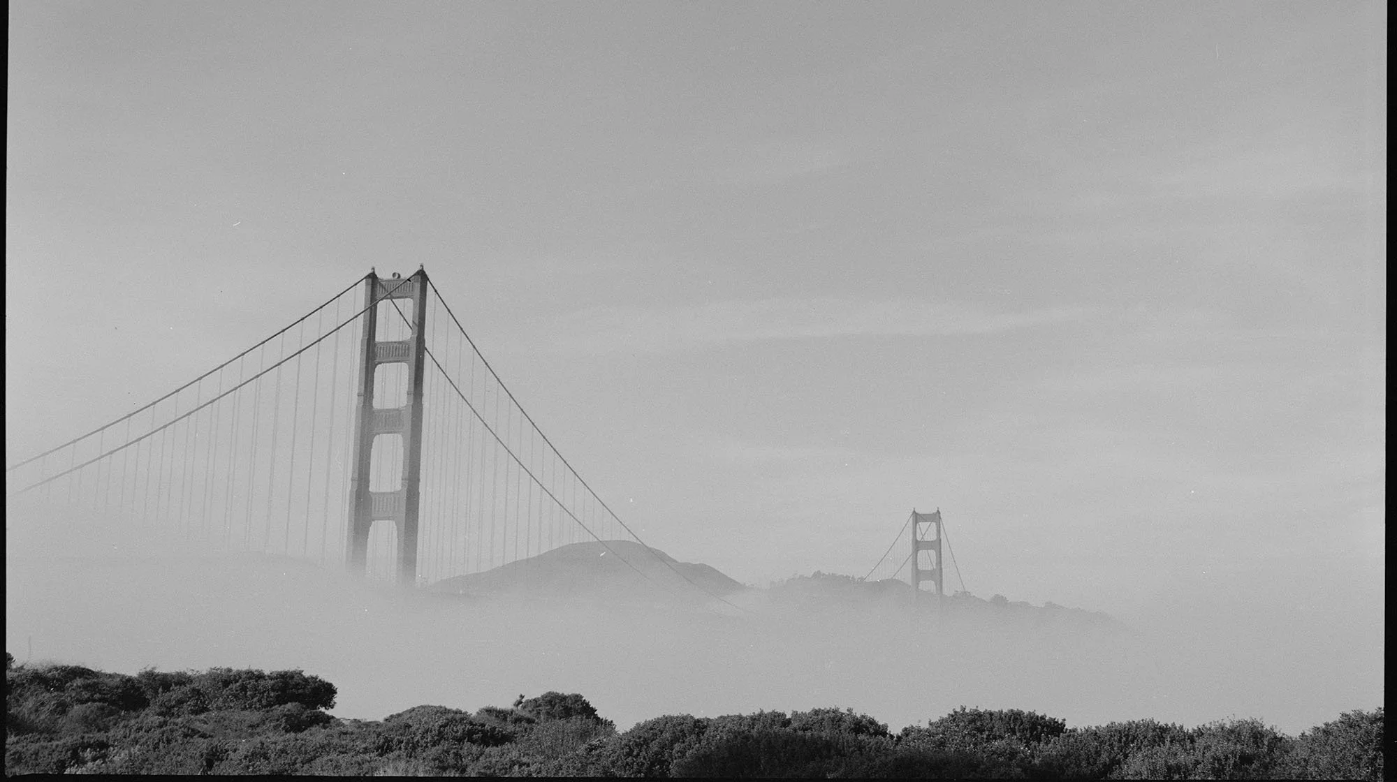 Black and white photograph of the Golden Gate Bridge with fog partially covering the lower part of the bridge and surrounding hills and trees.