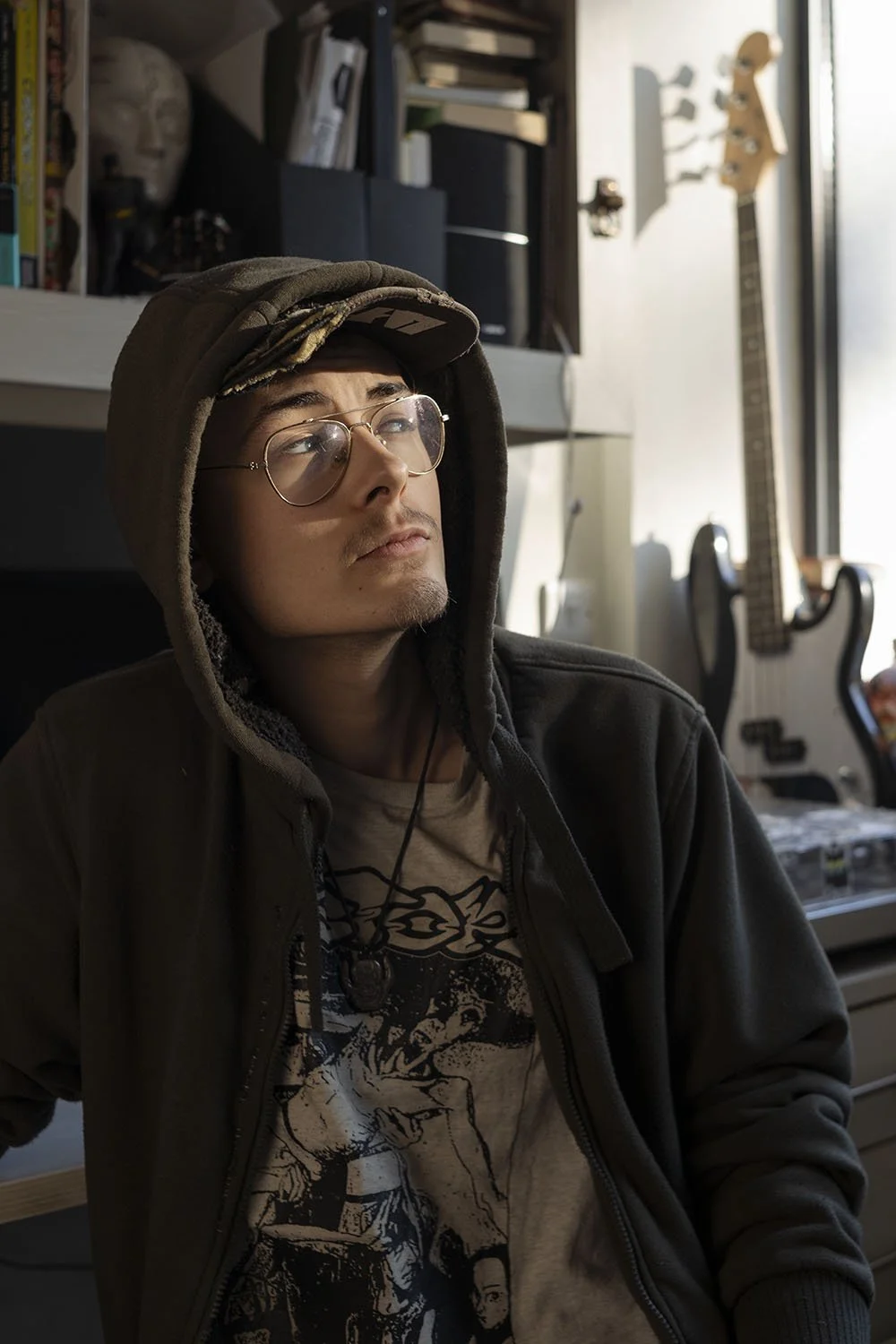 Young man with glasses wearing a hoodie and graphic T-shirt sitting in a room with musical instruments and shelves, looking contemplative.