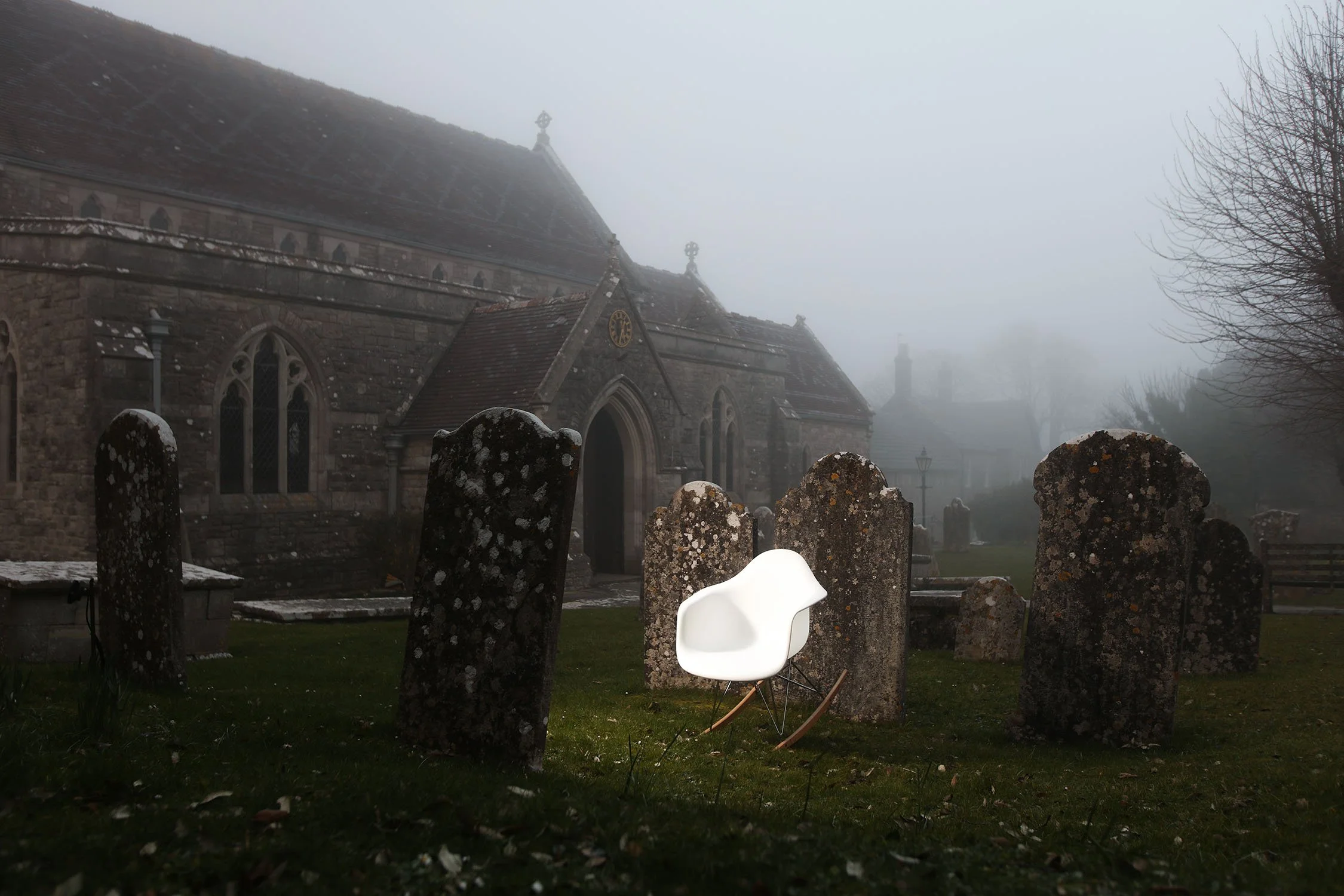 A British foggy graveyard with weathered tombstones, a church in the background, and barren trees, with a modern white chair in the foreground.