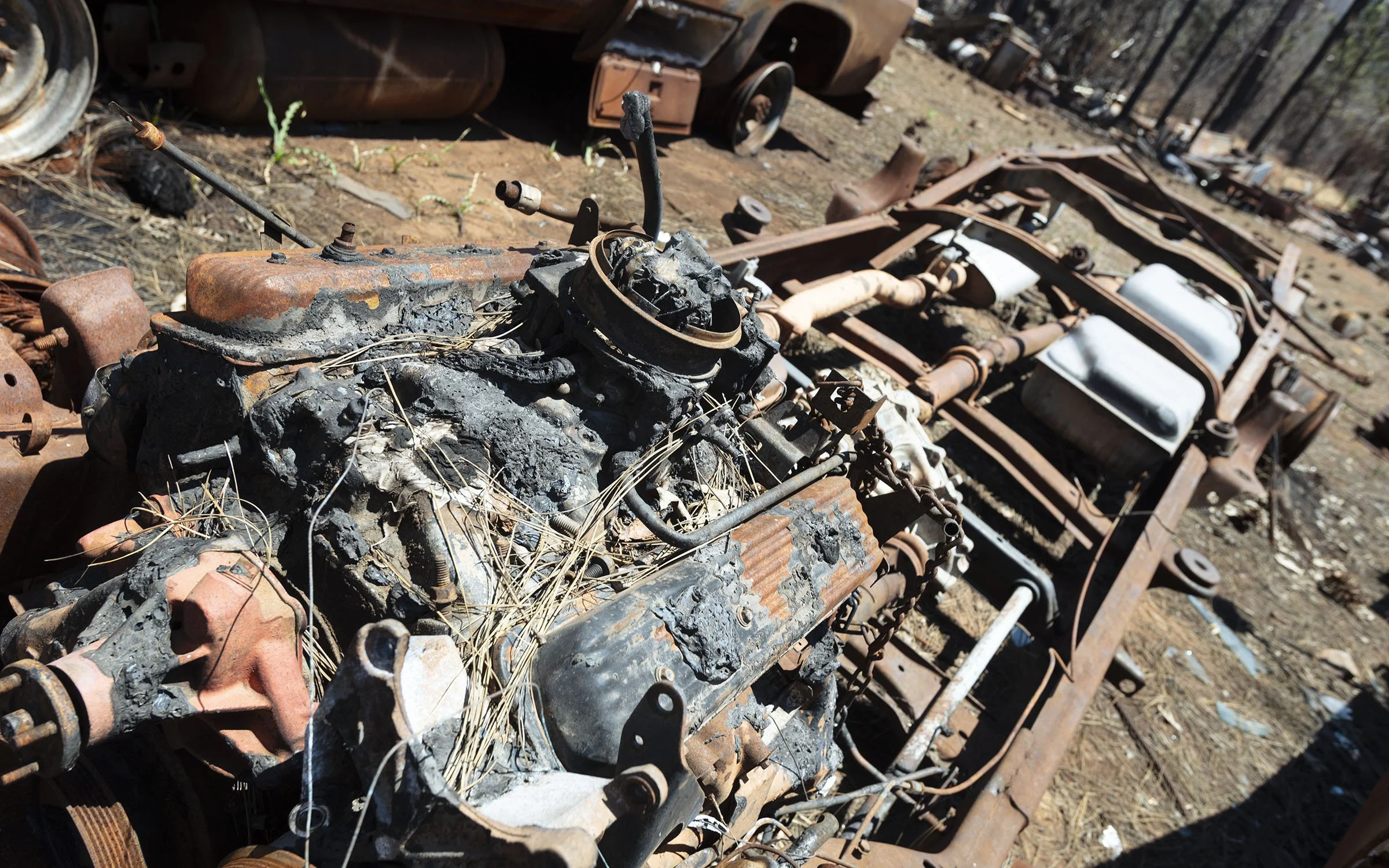 Burnt and rusted vehicle parts, with a destroyed engine in the foreground, lying on dirt with fallen leaves, in a scrapyard or abandoned area.