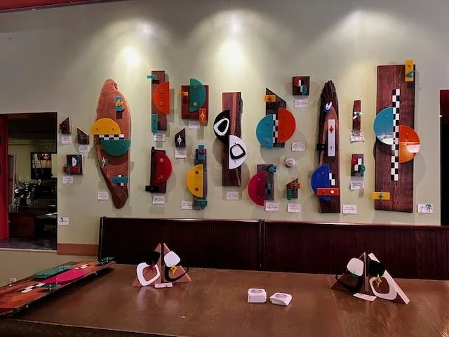Colorful geometric wooden art pieces on a beige wall, arranged artistically, with a wooden bench and tables in a well-lit room.
