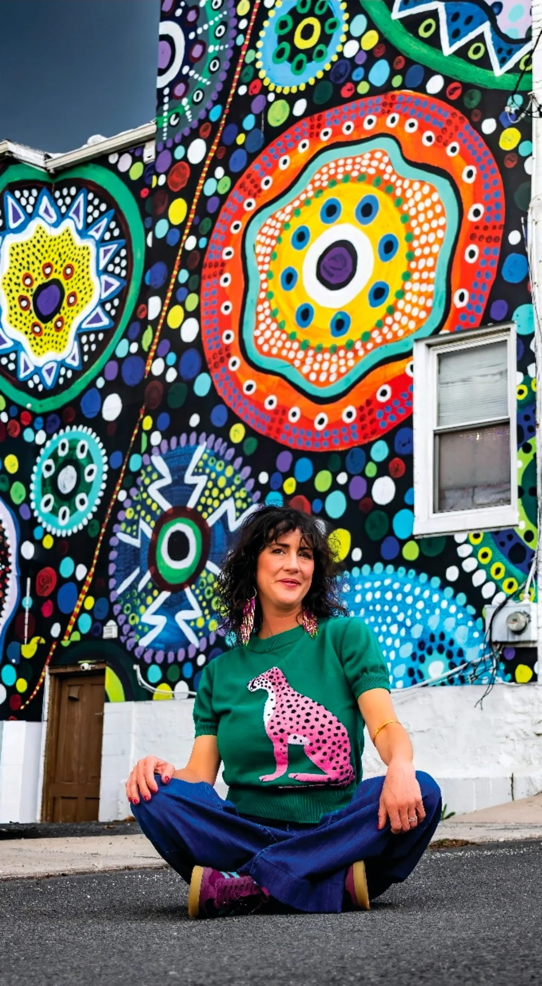 A woman with black curly hair and pink earrings sitting cross-legged on the street in front of a colorful, abstract mural with concentric circles and dots.