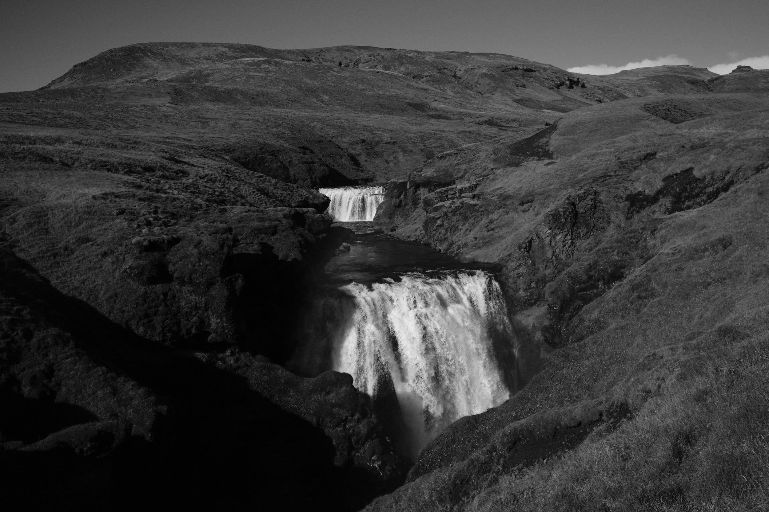Black and white photo of a landscape with a waterfall cascading down rocky terrain surrounded by rolling grassy hills.