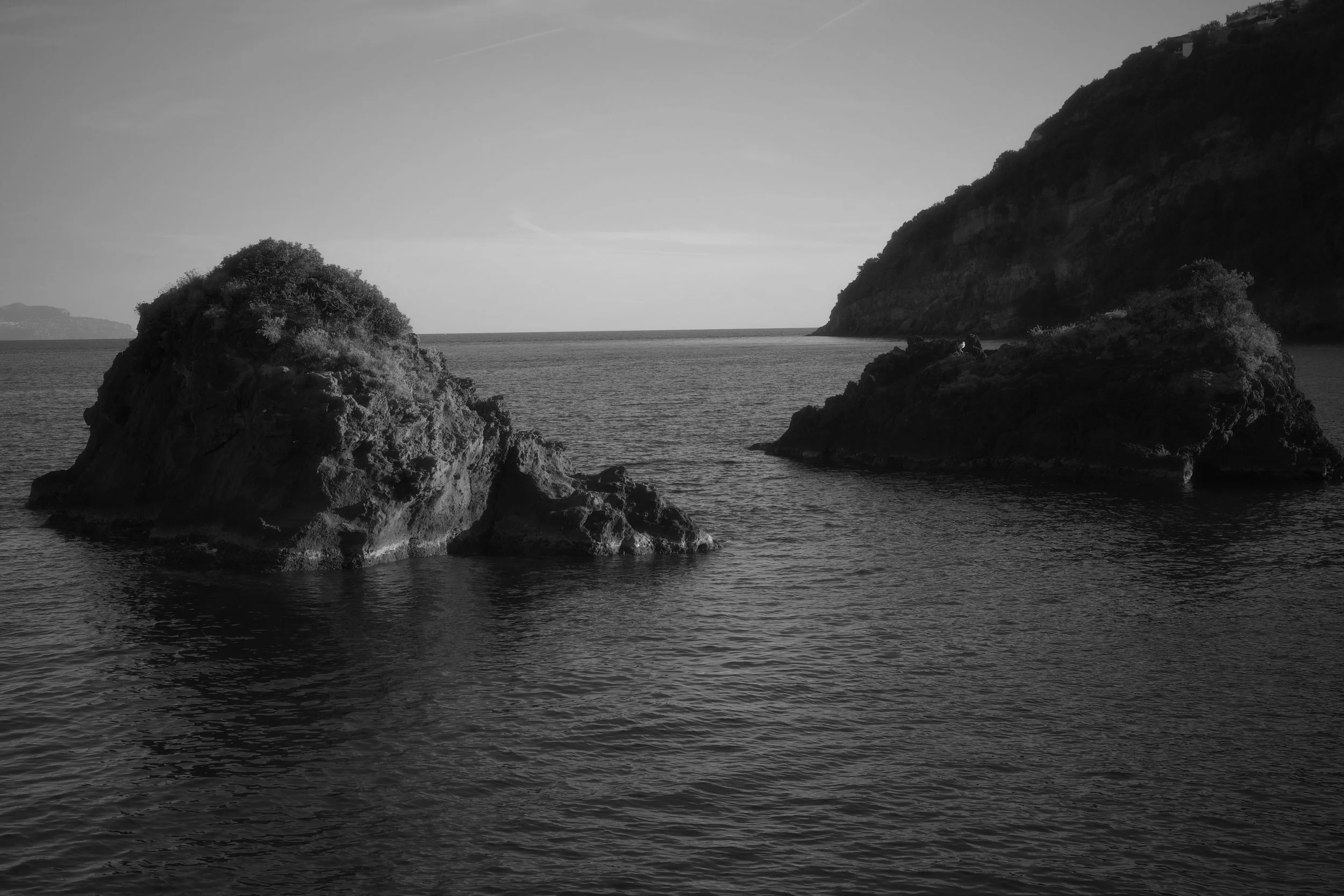 Black and white photograph of a seascape featuring two large rocks emerging from the water, with a hillside in the background.