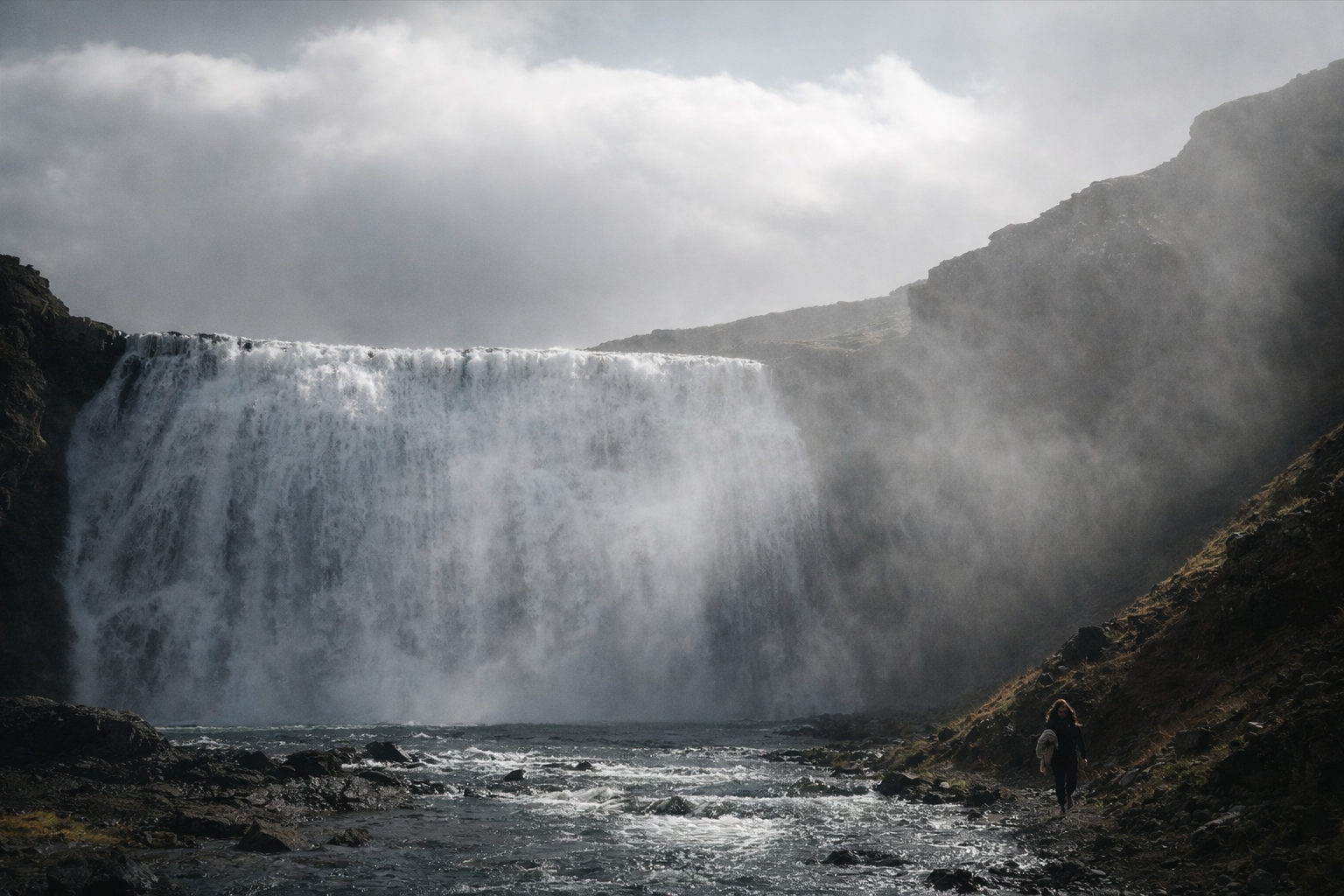 A person walking along a rocky riverbank near a large waterfall with mist rising and cloudy sky above.
