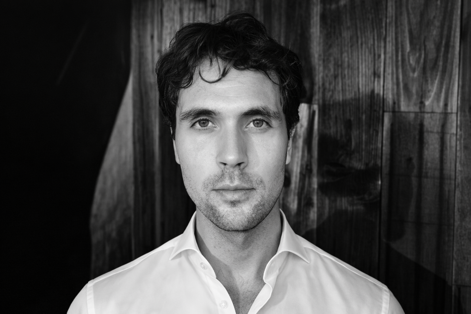 Black-and-white portrait of a man with dark, curly hair and light-colored shirt, standing against a wooden wall.
