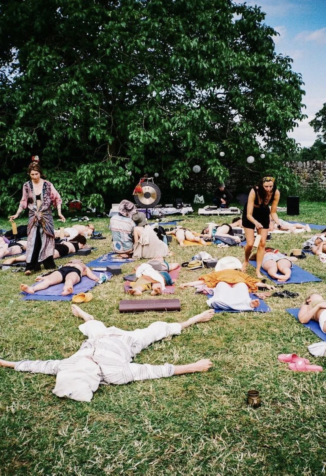 People practicing yoga outdoors on mats under a large tree in a park, with a musician playing gong and percussion instruments in the background.